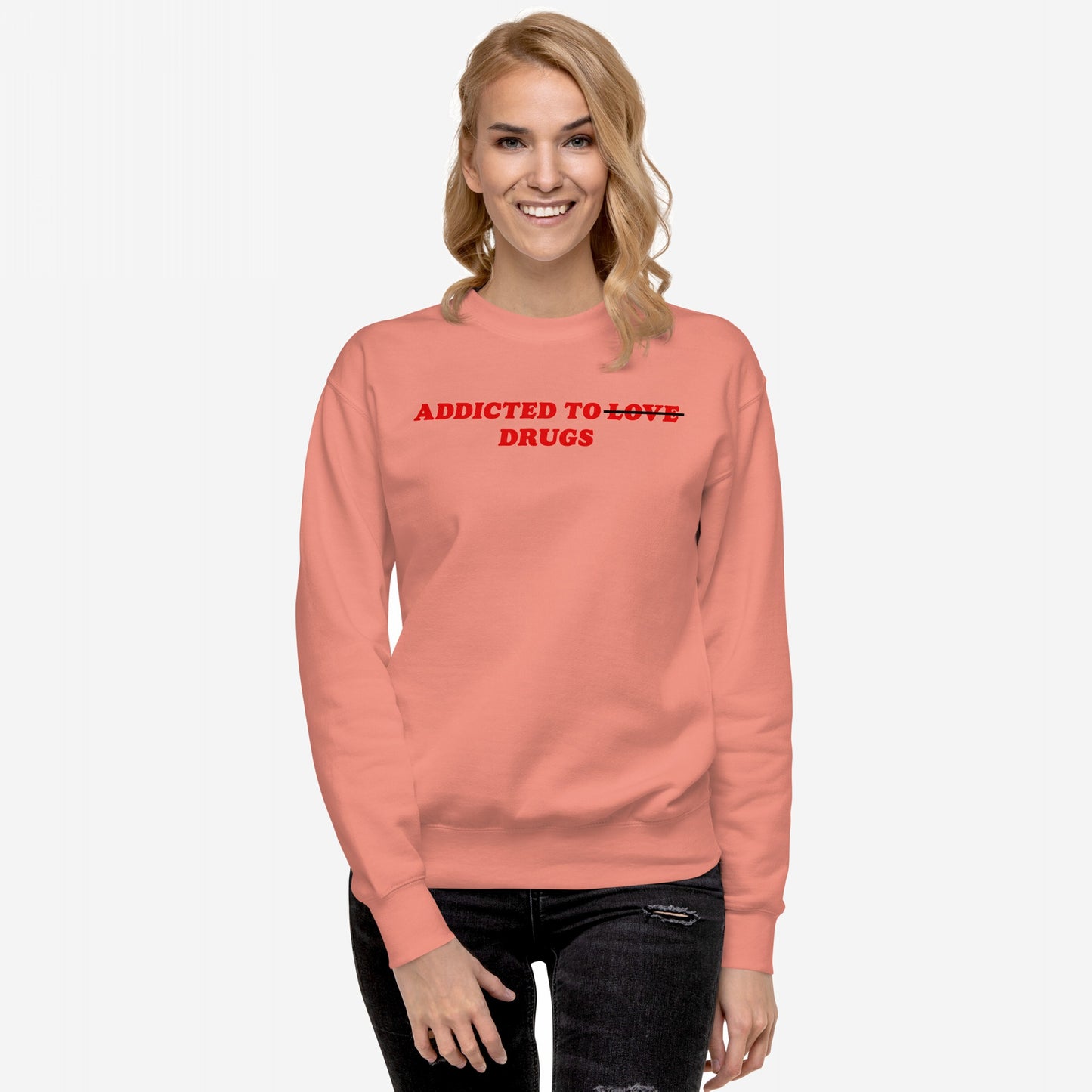 Young woman models the Addicted To Love / Drugs Hoodie & Sweatshirt, featuring bold text Addicted To ~~Love~~ Drugs on a coral sweatshirt, symbolizing personal struggle and self-awareness. She's styled with distressed jeans.