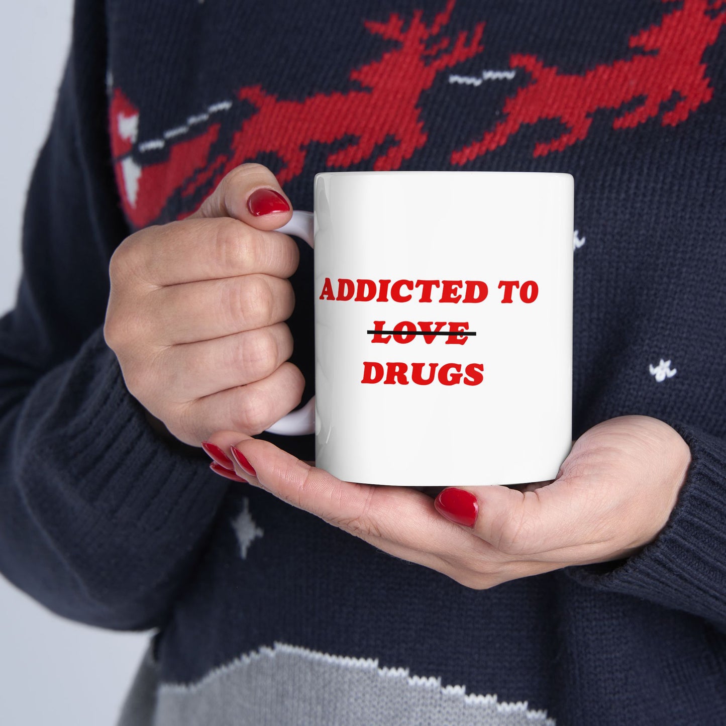 Hands with red nails hold the Addicted To Love / Drugs Mug, featuring bold red text with a humorous strikethrough design. The person wears a dark blue sweater with a red reindeer pattern.
