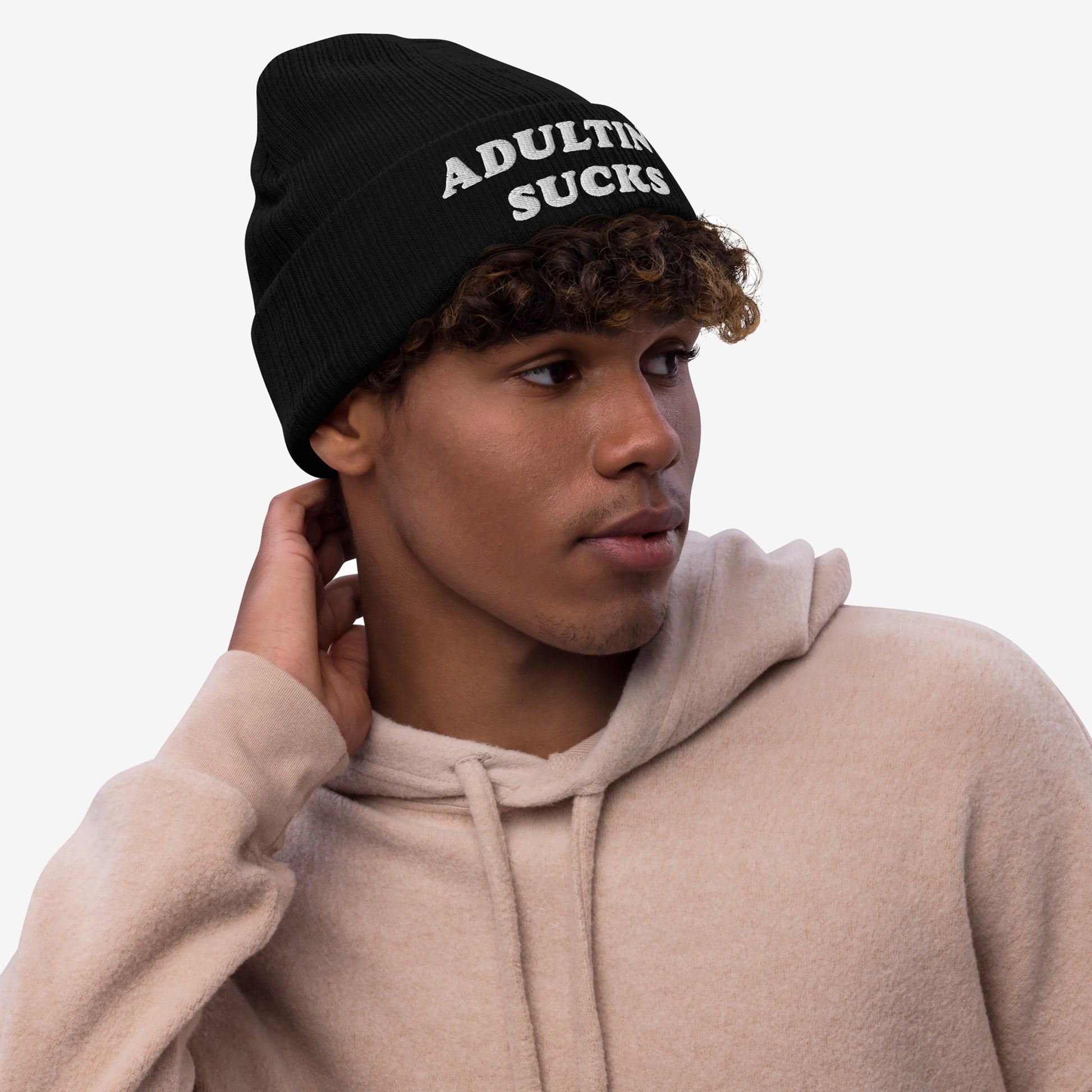 Young man in a light beige hoodie wears the Adulting Sucks hat, a black knit beanie with bold embroidery, capturing a relatable and stylish sentiment ideal for students and tired professionals.
