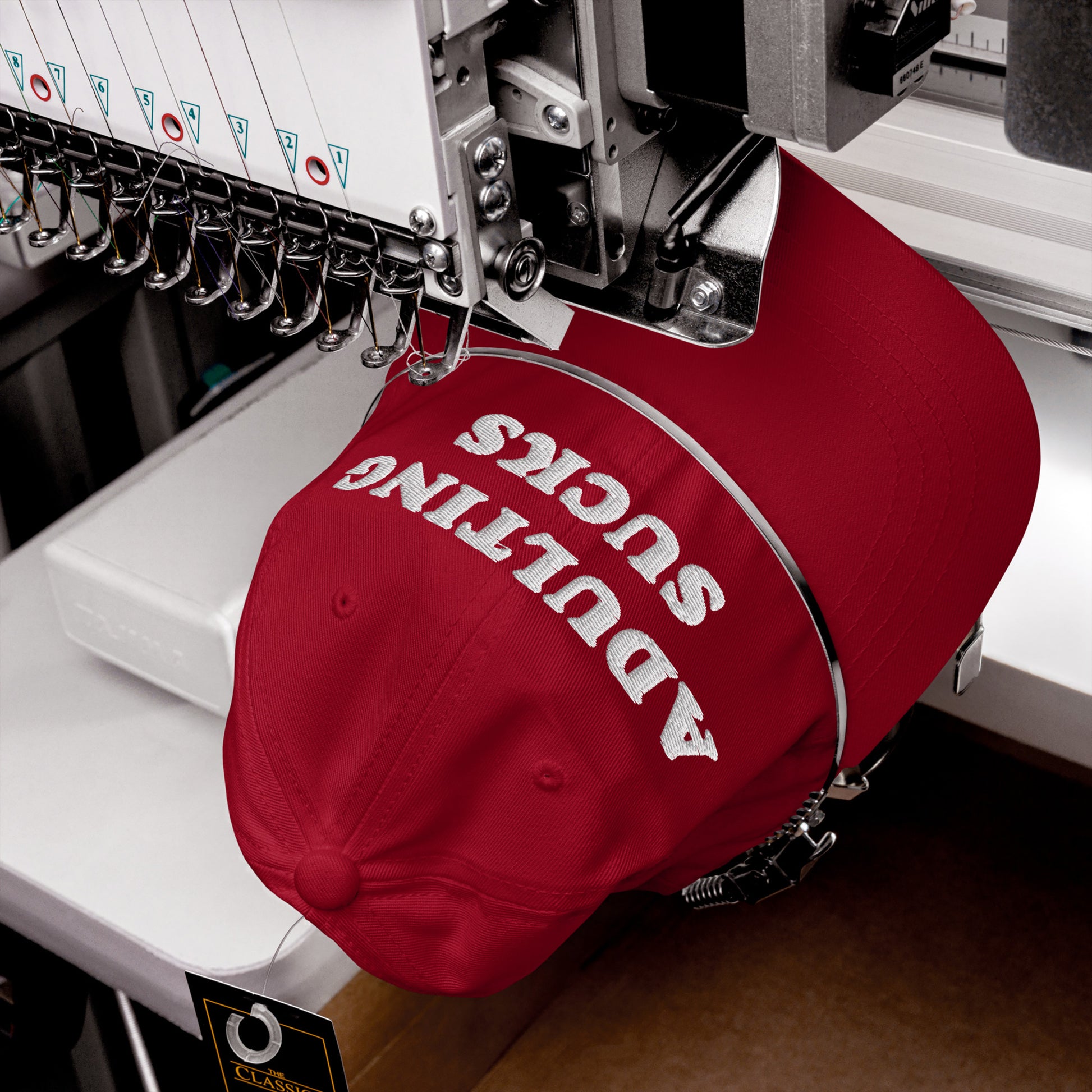 A red baseball-style cap being embroidered with ADULTING SUCKS on a multi-needle machine, capturing a bold statement ideal for those opposing adult life's pressures.