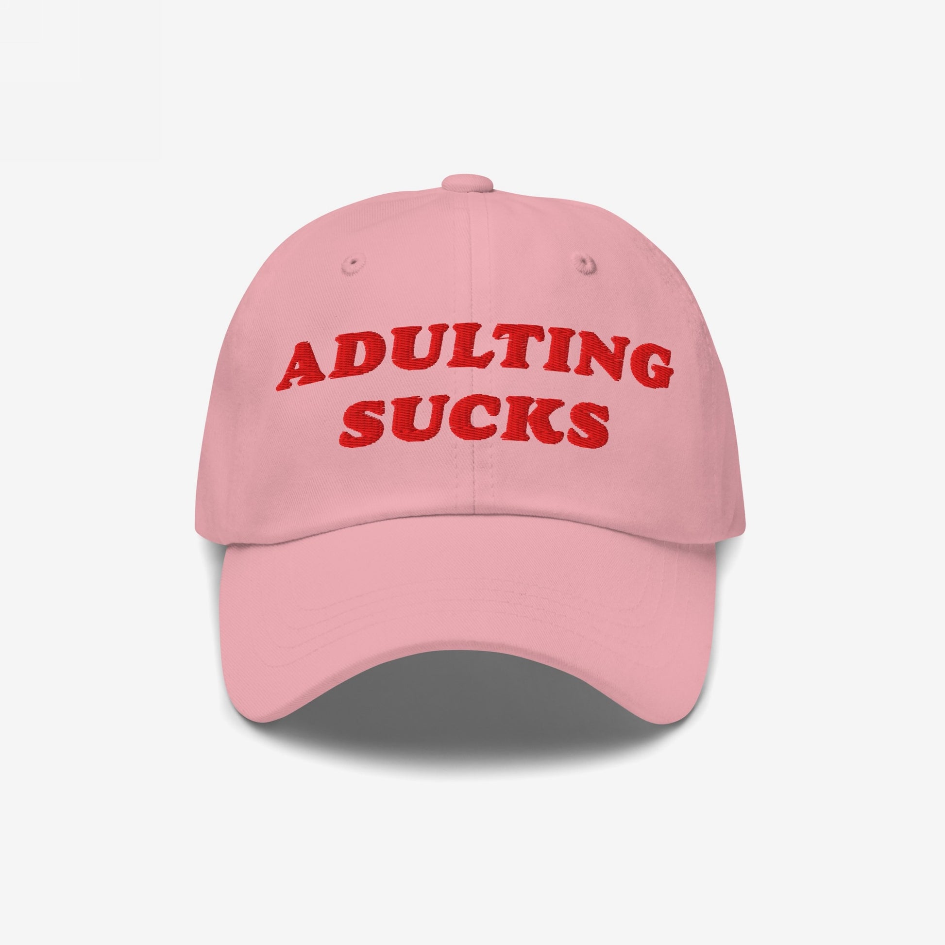 Pink baseball-style cap with bold red embroidery reading ADULTING SUCKS, highlighting a playful, relatable message for those overwhelmed by adult responsibilities. Perfect for casual wear and expressing individuality.