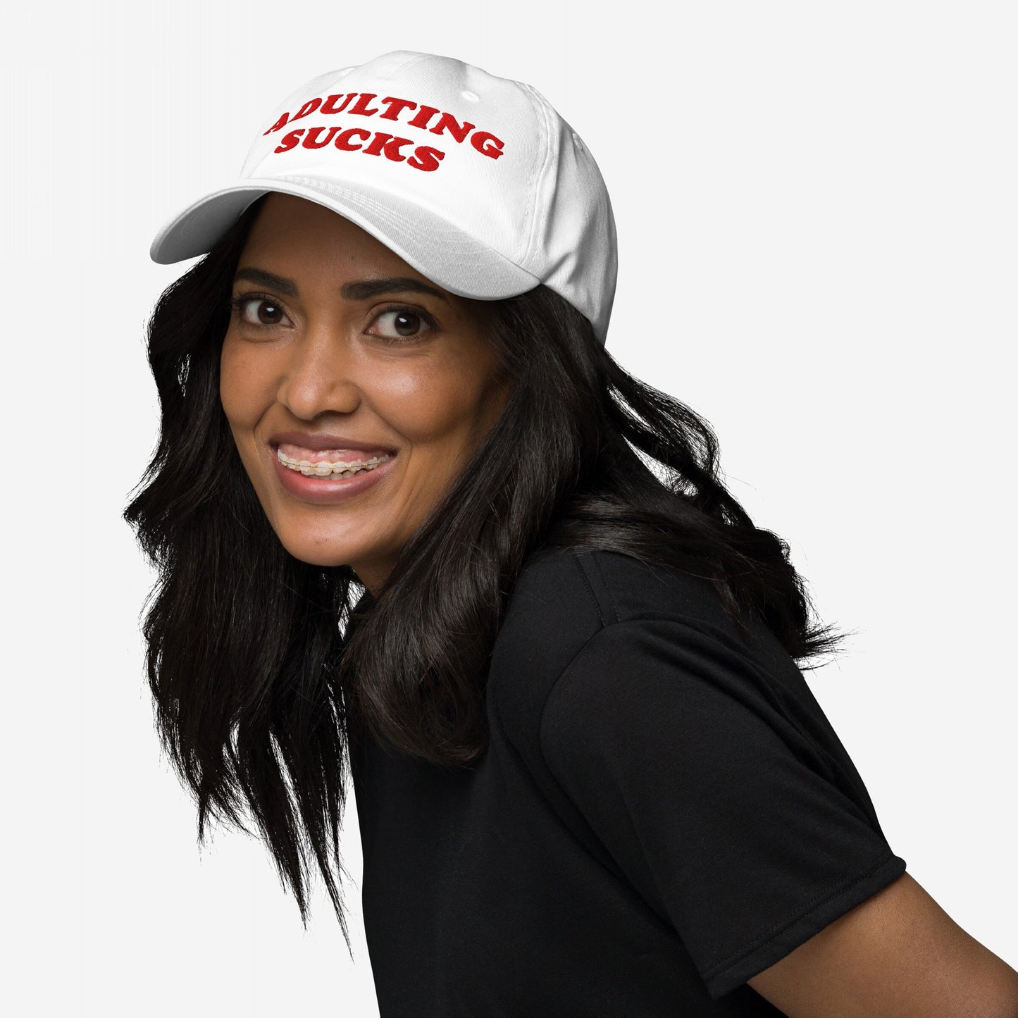 Young person with long dark hair smiles, wearing an Adulting Sucks hat with red embroidery. The white cap humorously conveys the struggles of adulthood, embodying a candid yet playful message.