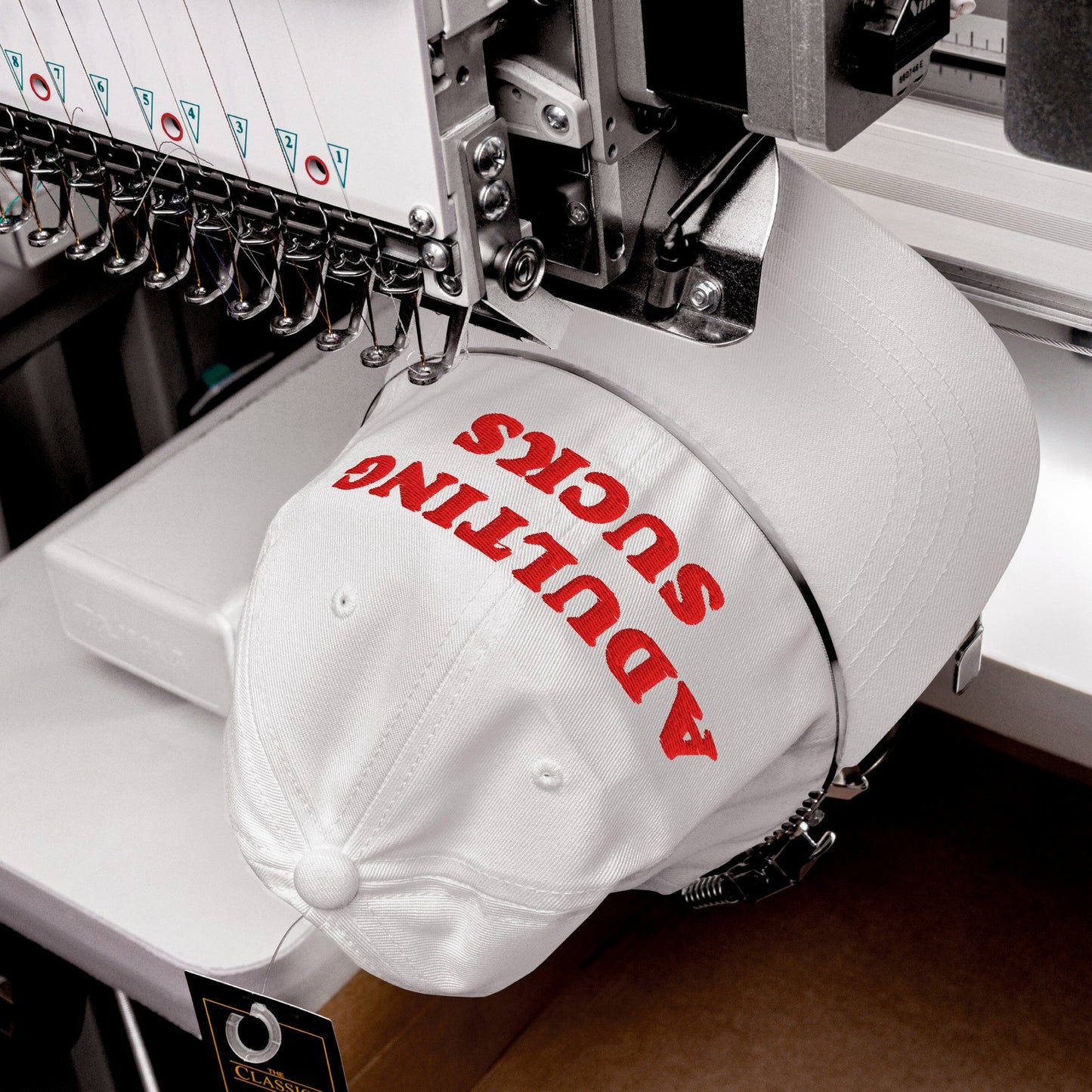 The Adulting Sucks Hat is shown being embroidered in red thread on a machine, capturing its bold message that resonates with those feeling overwhelmed by life's challenges.
