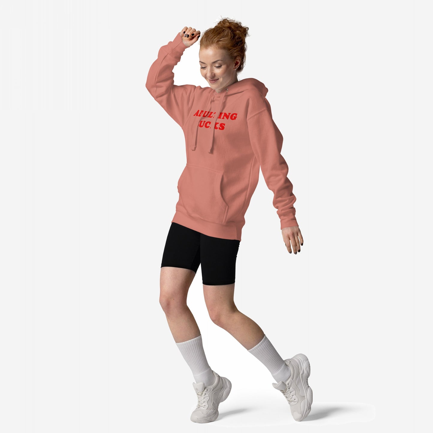 Young woman playfully poses in the Adulting Sucks Hoodie & Sweatshirt, showcasing its bold statement print. She's styled with bike shorts, socks, and sneakers, embodying a carefree, relatable vibe.