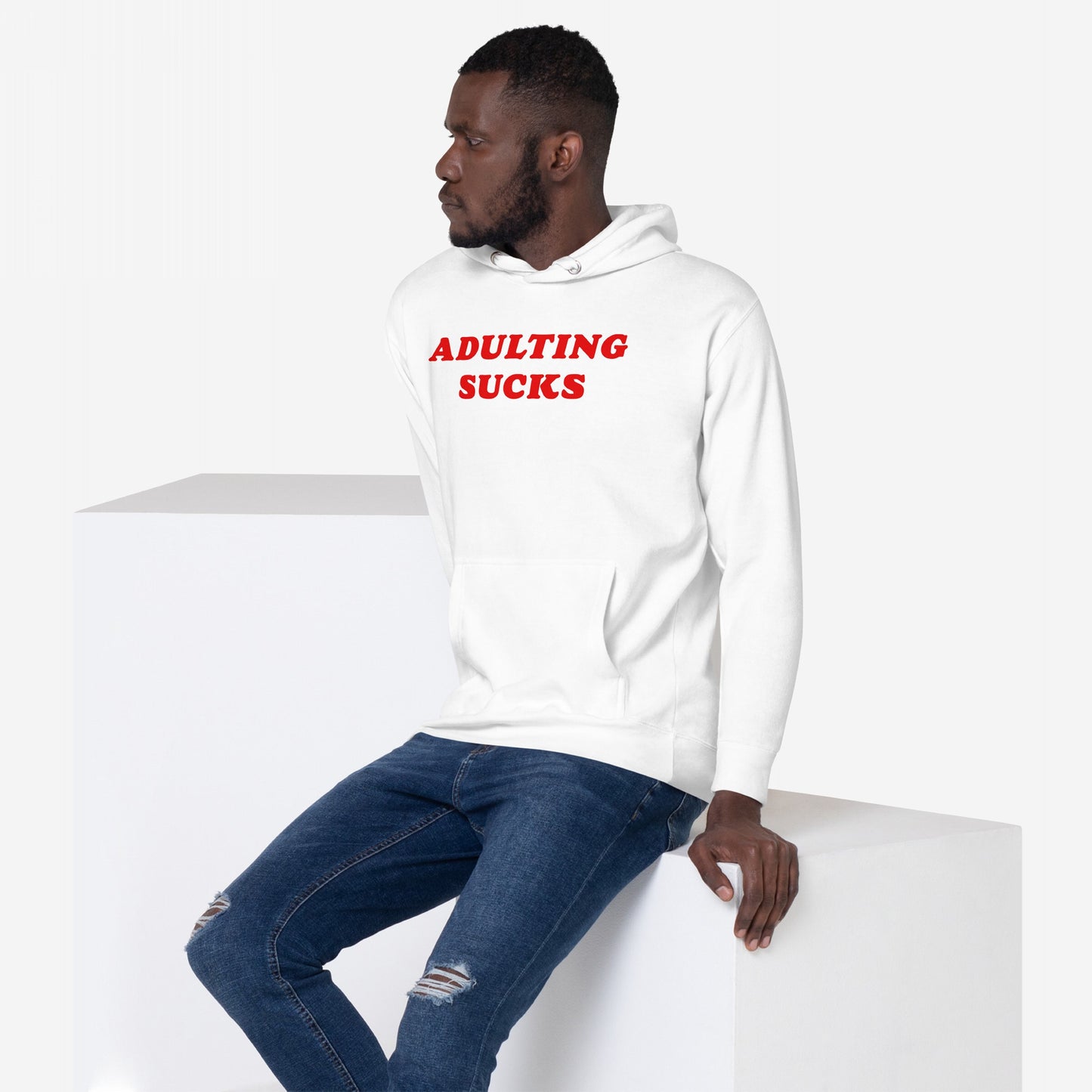 Young man wears the Adulting Sucks Hoodie & Sweatshirt with bold red text, expressing humor and relatability. Paired with ripped jeans, it embodies casual defiance against daily challenges.