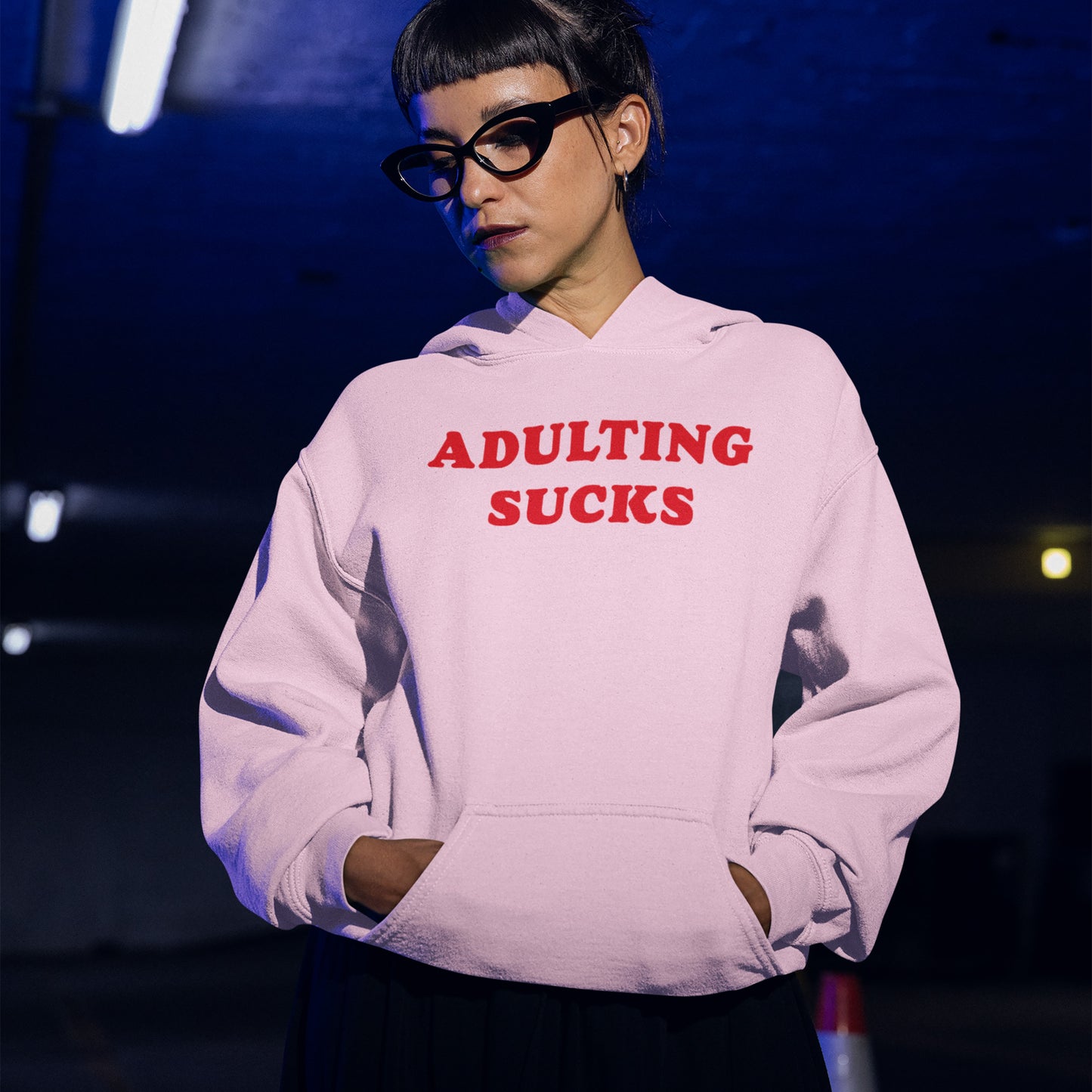 Person wearing a light-colored Adulting Sucks hoodie with bold red lettering, standing in a dimly lit indoor space, embodying a humorous, relatable vibe ideal for casual comfort and self-expression.