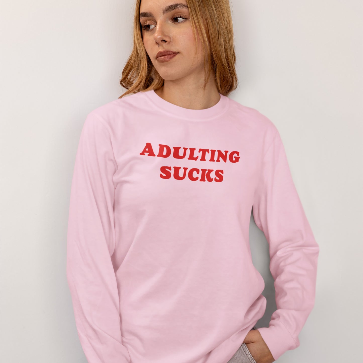Young person wearing the Adulting Sucks T-shirt, featuring a bold red slogan on a pastel-pink long-sleeve shirt. The design combines retro charm and humor, ideal for expressing modern frustrations.