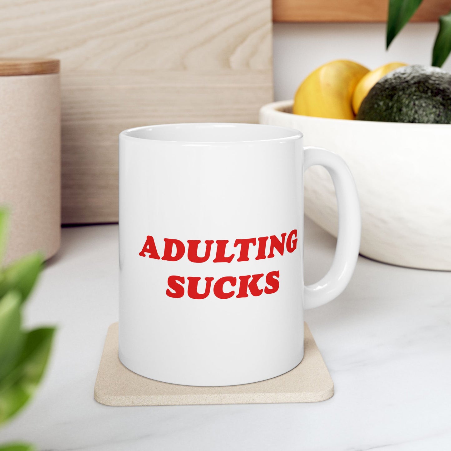 White ceramic Adulting Sucks mug on cork coaster, boldly stating its message in red, embodying a rebellious spirit. Set on a countertop with subtle background elements. Ideal for those challenging societal norms.