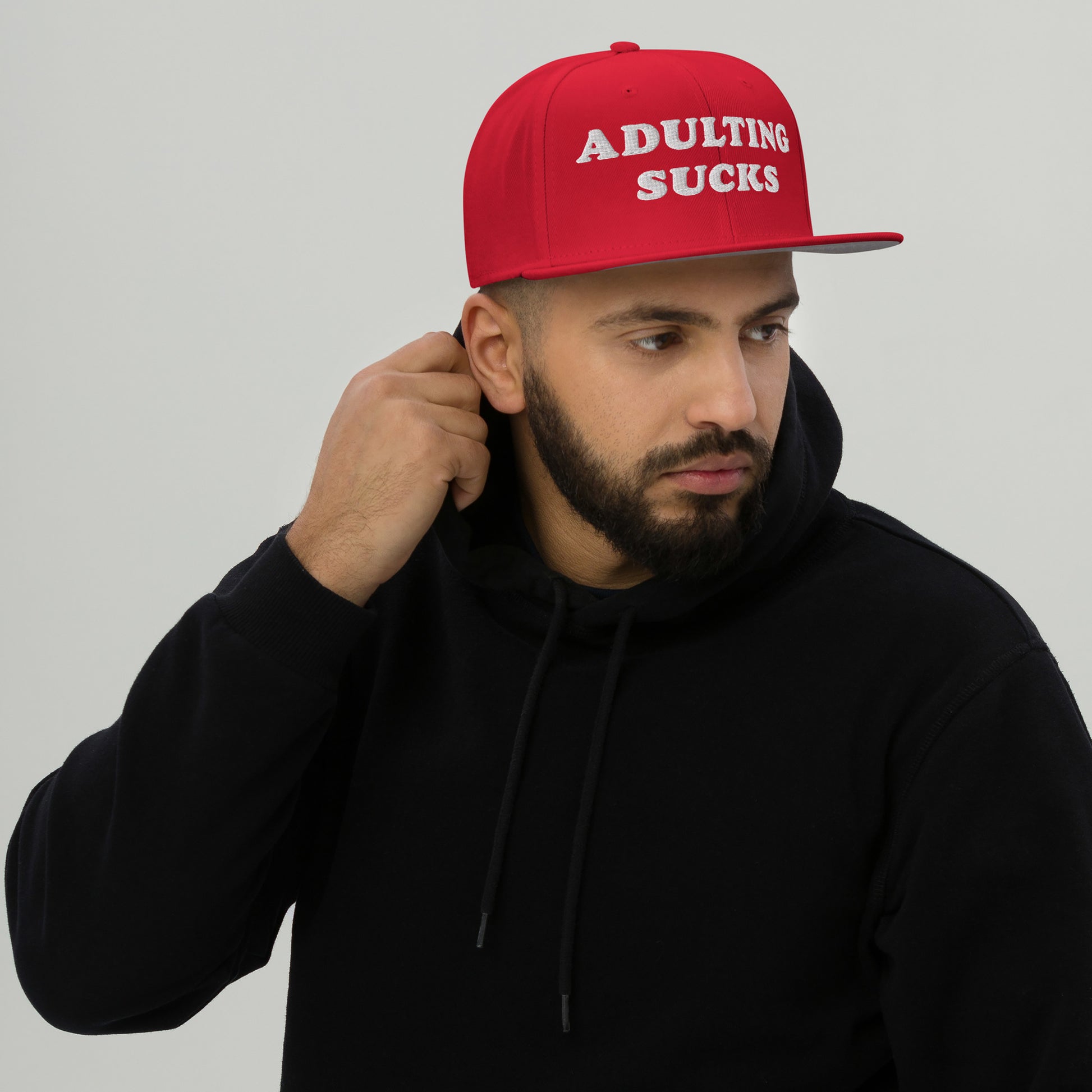 Man wearing a black hoodie and a red Adulting Sucks cap, expressing frustration. The bold, embroidered message captures the hat's playful critique of adult responsibilities, resonating with millennial and Gen Z wearers.