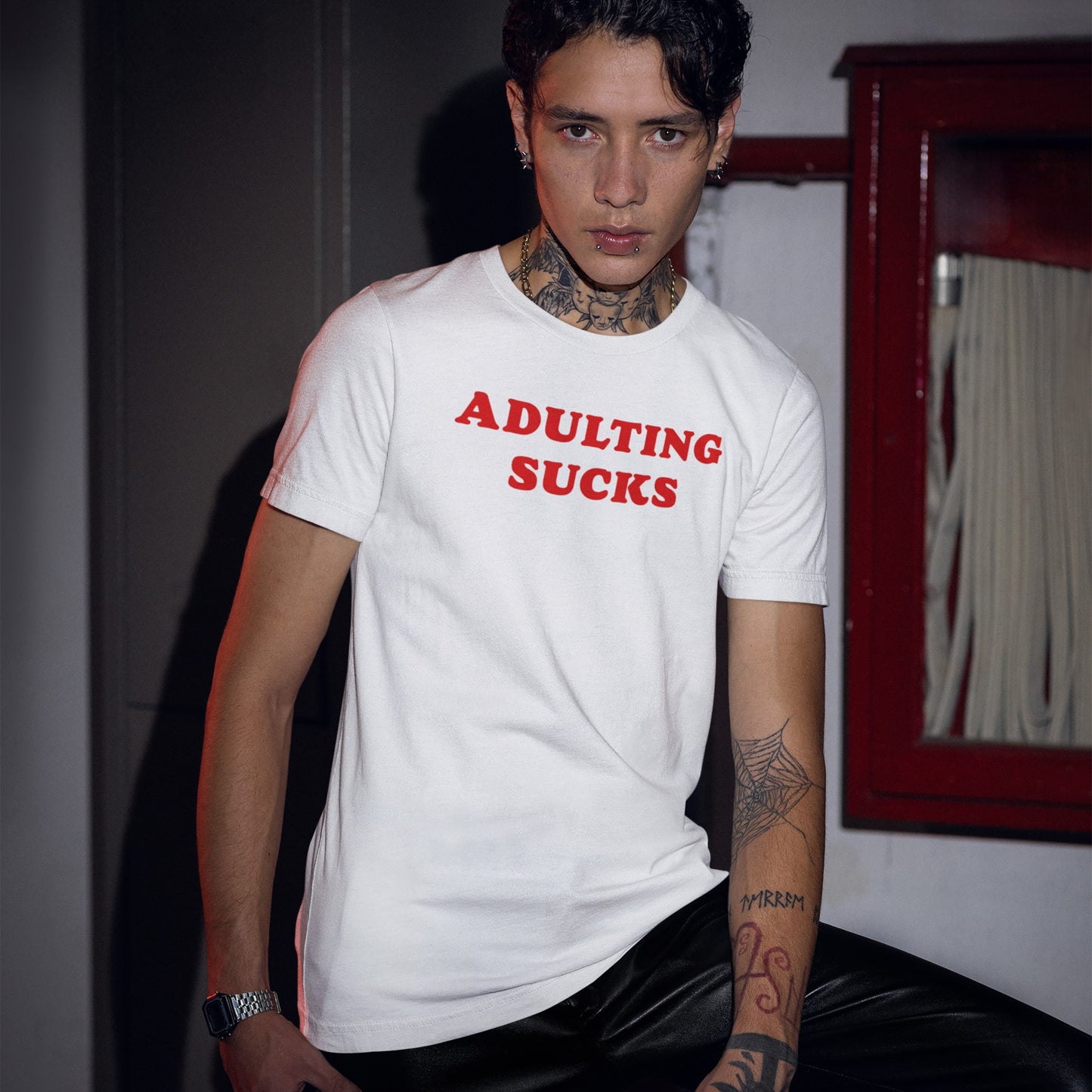 Adulting Sucks T-shirt with bold red text on a young person, paired with black pants, tattoos, and piercings, embodying a rebellious, authentic style in an industrial setting.