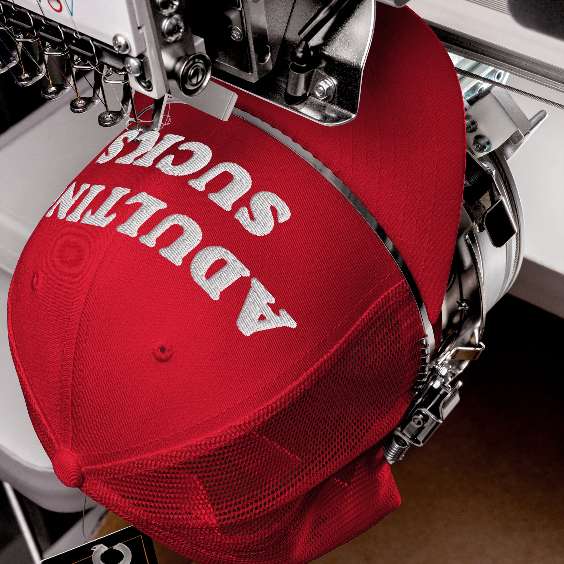 Close-up of an embroidery machine stitching “ADULTING SUCKS” on a red cap, showcasing a bold statement hat ideal for expressing relatable frustration with adult responsibilities.