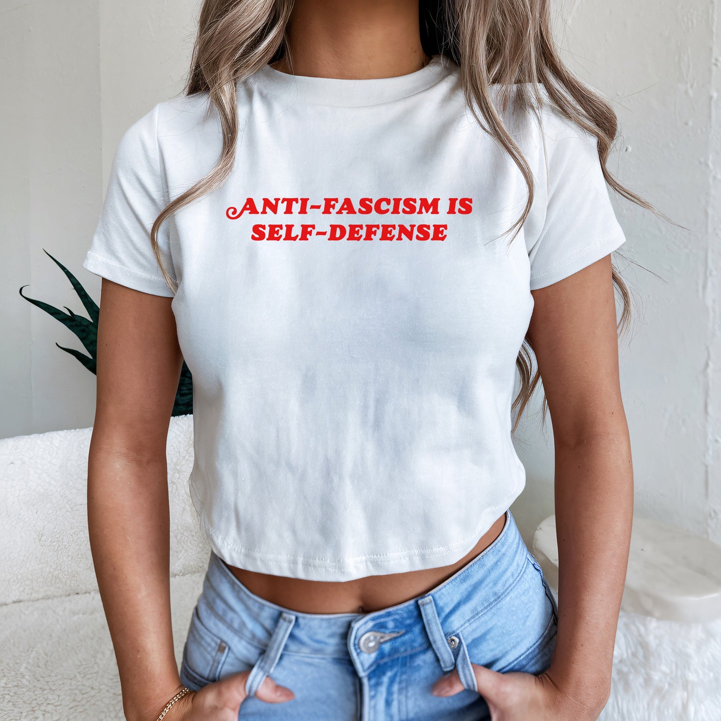 Person wearing the Anti-Fascism Is Self-Defense Women's Baby Tee, featuring bold red lettering on a fitted white crop-style T-shirt. Paired with light-wash jeans, embodying a stance of resistance and equality.