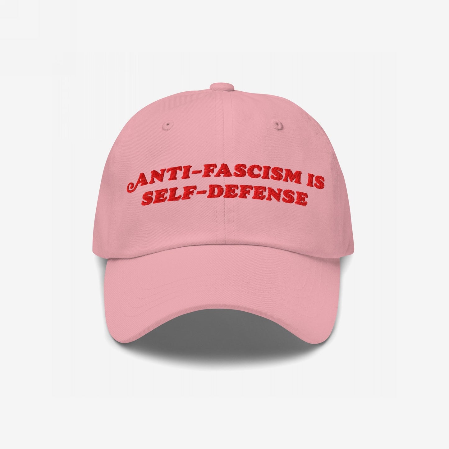 Light pink baseball cap with the slogan Anti-Fascism Is Self-Defense embroidered in red across the front, featuring a curved brim and classic six-panel design.