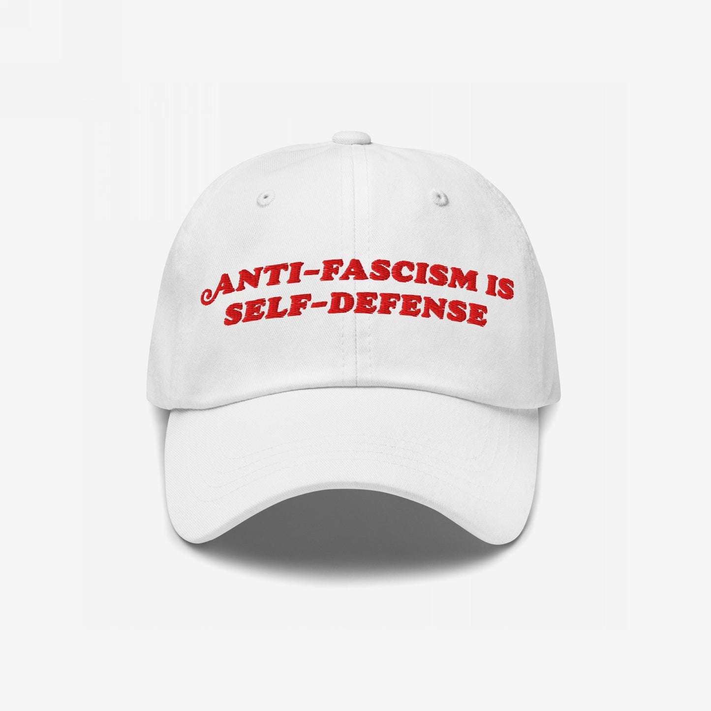 White baseball-style cap with red embroidered text ANTI-FASCISM IS SELF-DEFENSE, conveying a bold message of resistance. Perfect for advocates of equality and change, this durable, statement accessory supports Amnesty International.