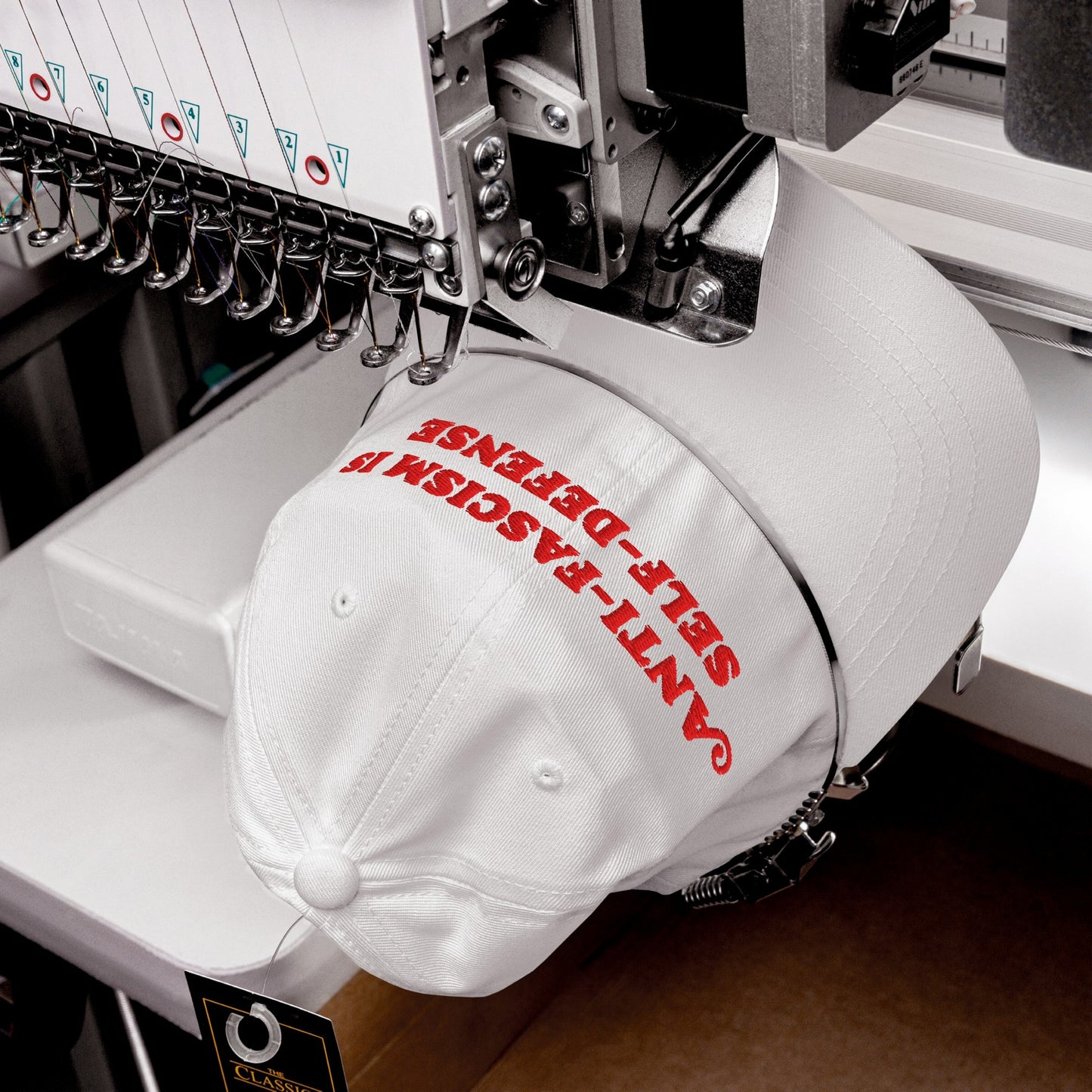 Multi-needle embroidery machine stitching “ANTI-FASCISM IS SELF-DEFENSE” on white cap, showcasing the Anti-Fascism Is Self-Defense Hat. Needles and thread guides visible, emphasizing resistance-themed fashion. Classic swing tag attached.