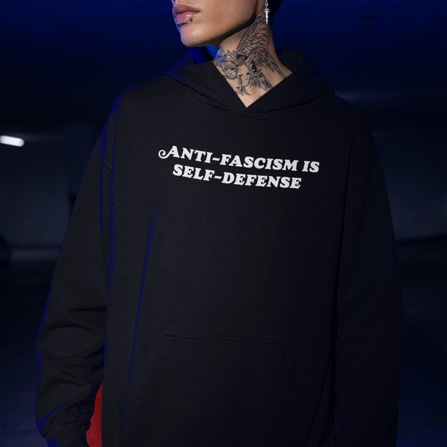Person wearing black hoodie with ANTI-FASCISM IS SELF-DEFENSE in bold white letters, neck tattoo, lip piercing, and earring visible, embodying activism and justice through the Anti-Fascism Is Self Defense Shirt.