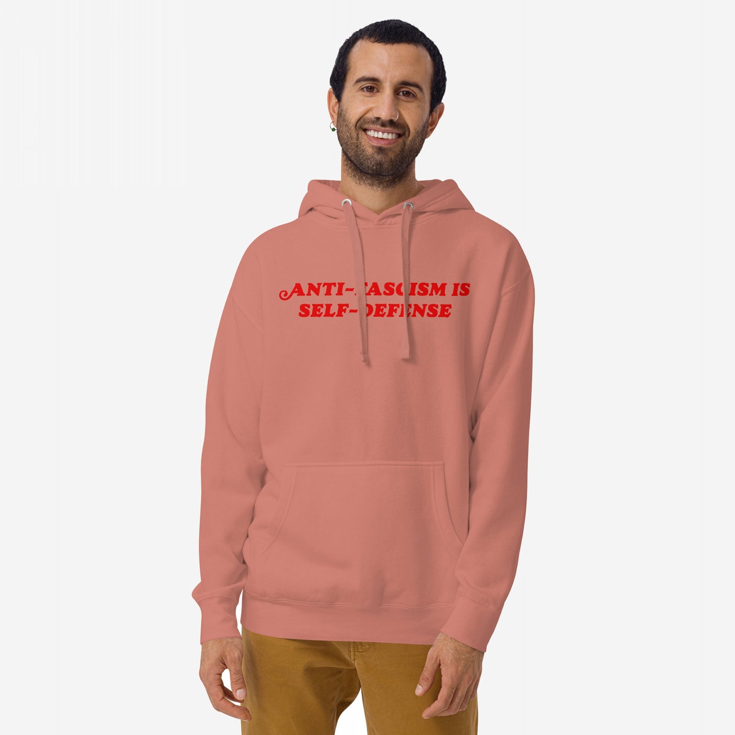 Smiling man models the Anti-Fascism Is Self-Defense Hoodie, featuring bold, impactful messaging on a muted red backdrop, embodying a stand against hate and promoting equality.