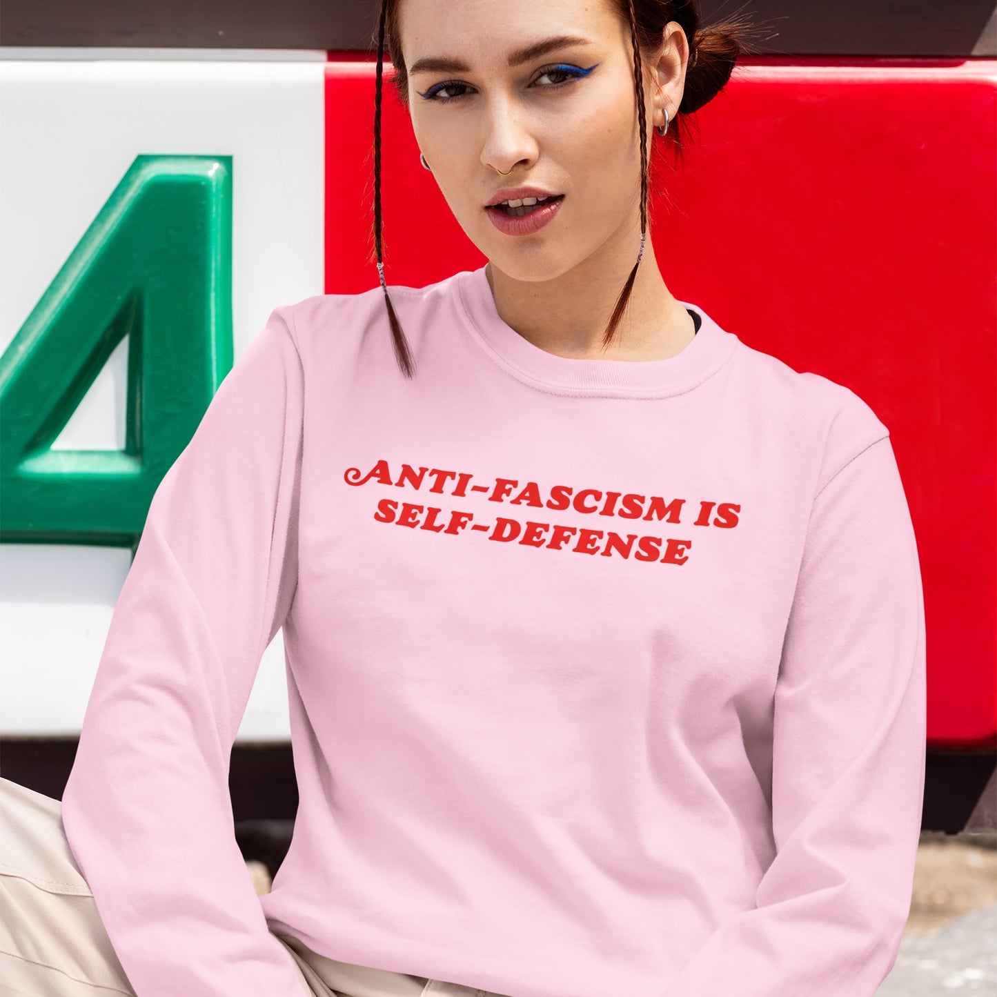 Young woman wearing the Anti-Fascism Is Self-Defense T-shirt, featuring bold red lettering on a pale-pink long sleeve, embodying resistance and activism, against a backdrop of broad green, white, and red bands.