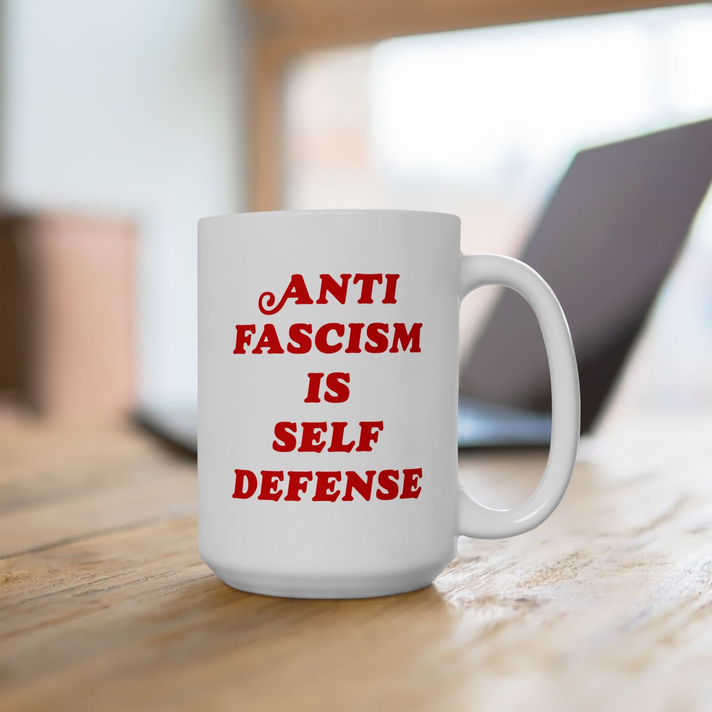White ceramic “Anti-Fascism Is Self-Defense” mug with bold red text, on a wooden desk, against a blurred office backdrop. Advocates resistance; ideal for those opposing hate and supporting equality.
