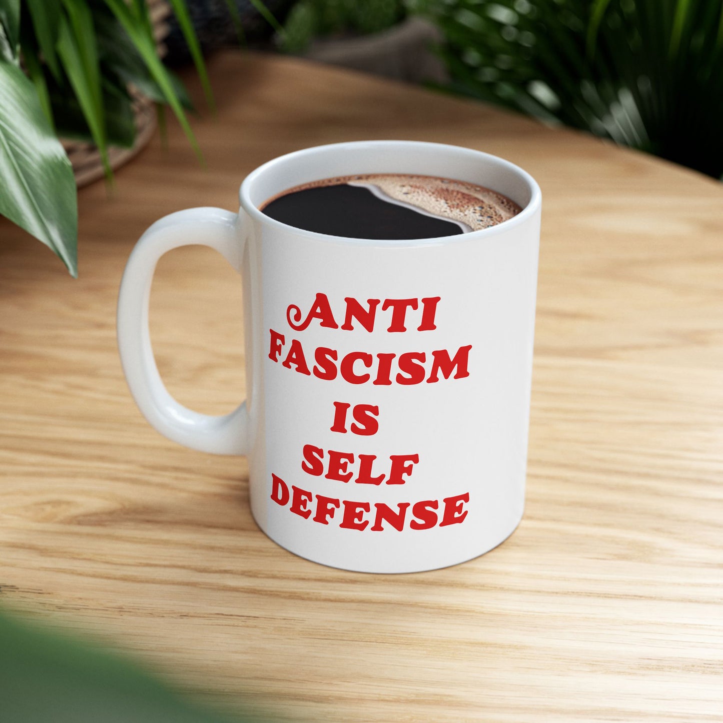 A white ceramic Anti-Fascism Is Self-Defense mug with bold red lettering filled with black coffee on a light-wood tabletop, surrounded by green potted plants, embodying strong convictions against fascism.
