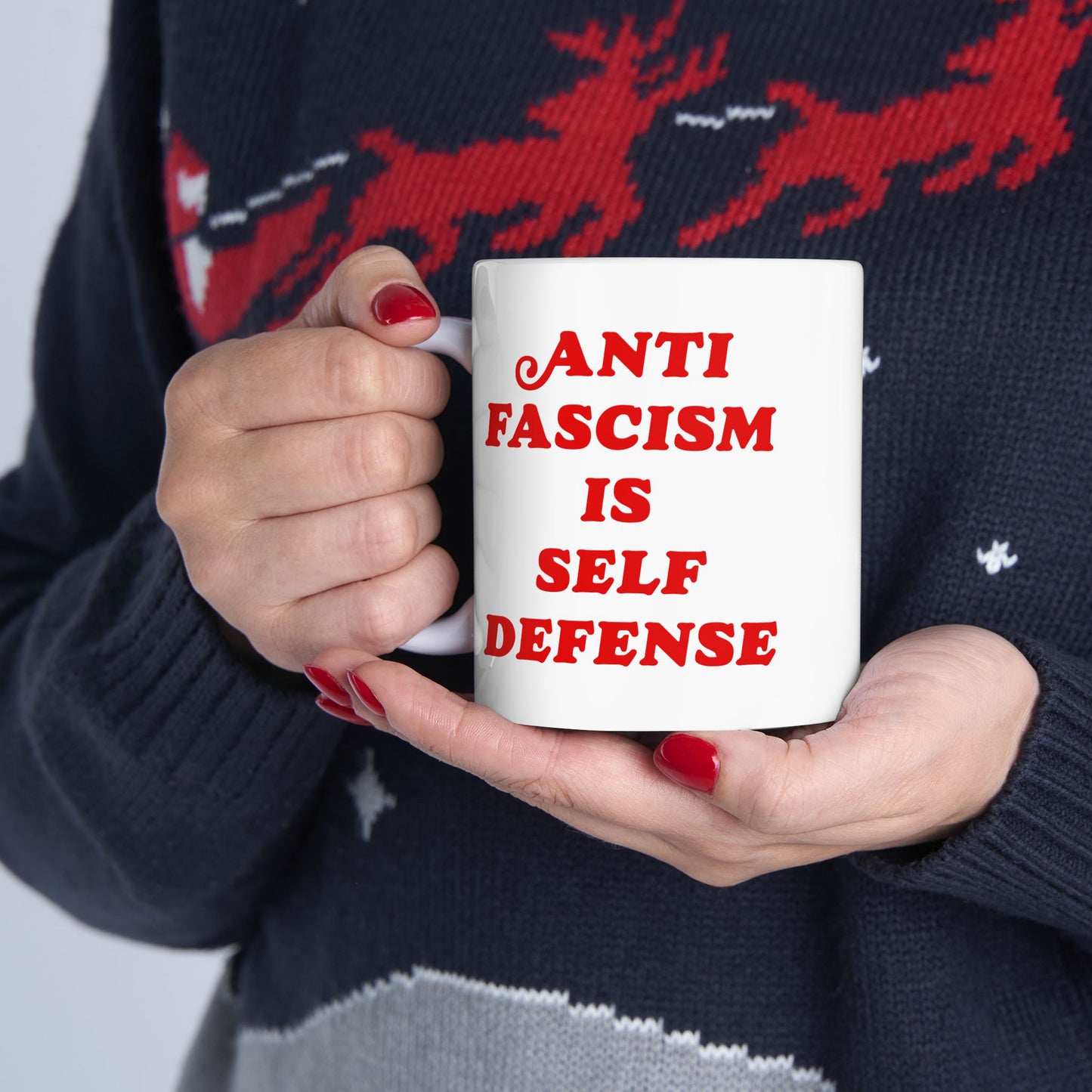 Person holding the Anti-Fascism Is Self-Defense Mug, featuring bold red text on a white ceramic surface. The mug is gripped with both hands, displaying red-painted fingernails and a dark sweater.