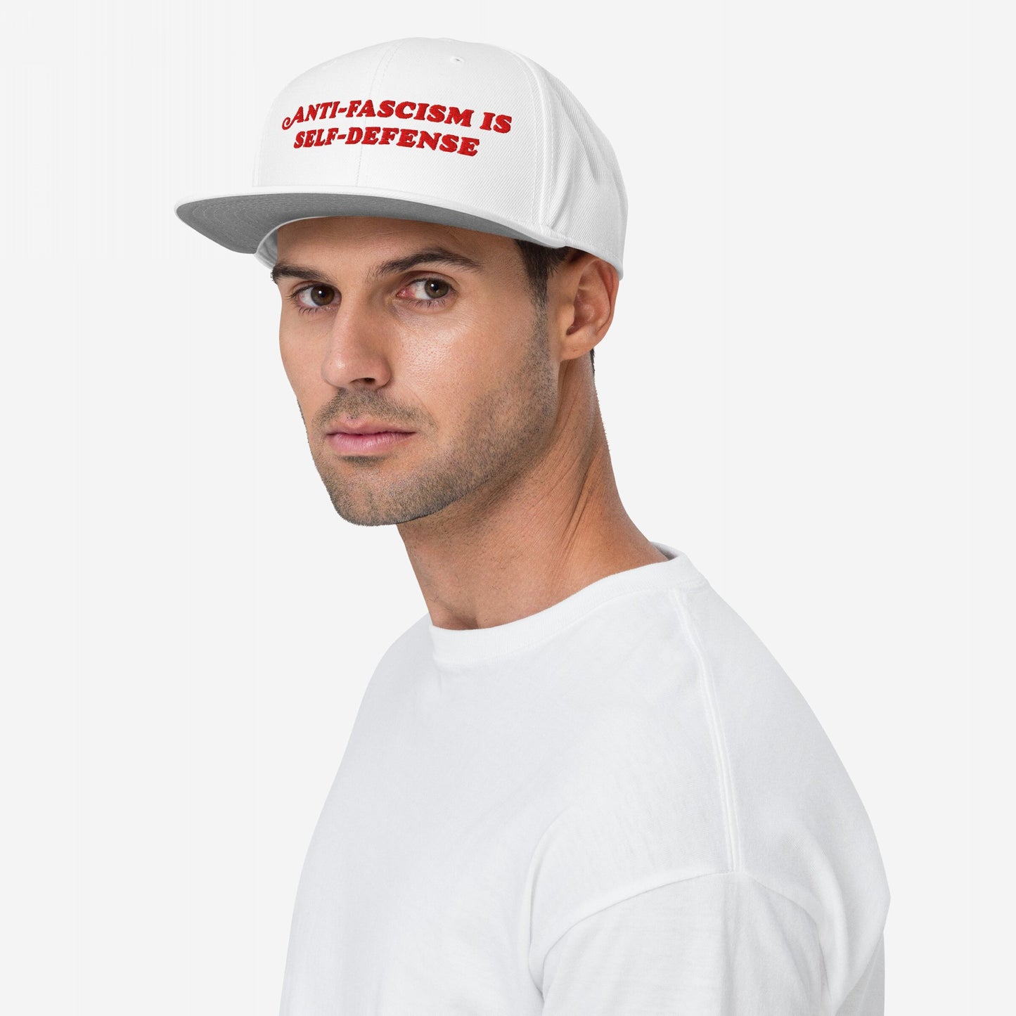 A young man wears the Anti-Fascism Is Self-Defense hat, showcasing bold red embroidered text on a white flat-bill cap, embodying fashion as resistance with a strong, clear message.