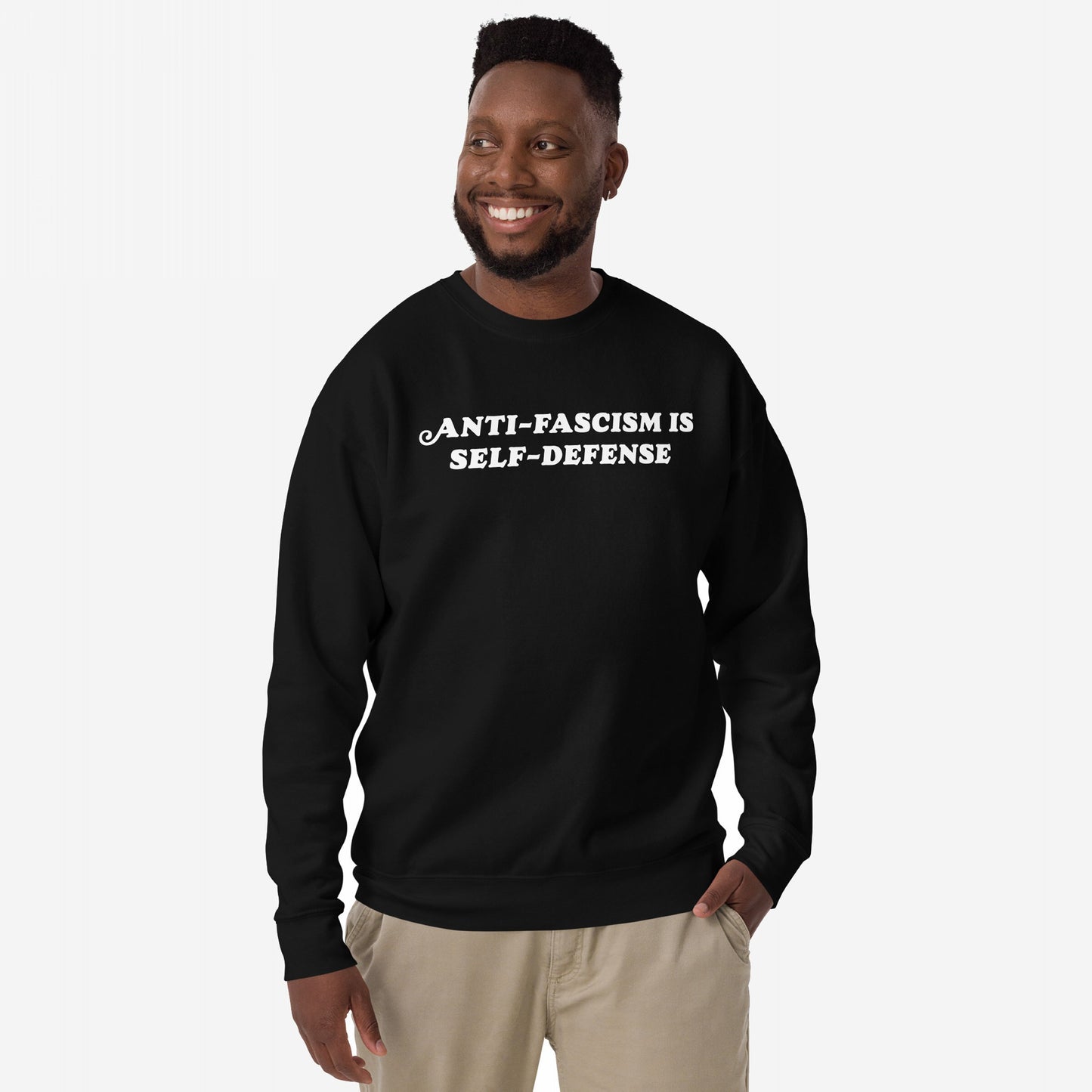 Man wearing the Anti-Fascism Is Self-Defense Hoodie & Sweatshirt, featuring bold white lettering on a black crewneck. He smiles confidently, embodying the garment's message of resistance and equality.