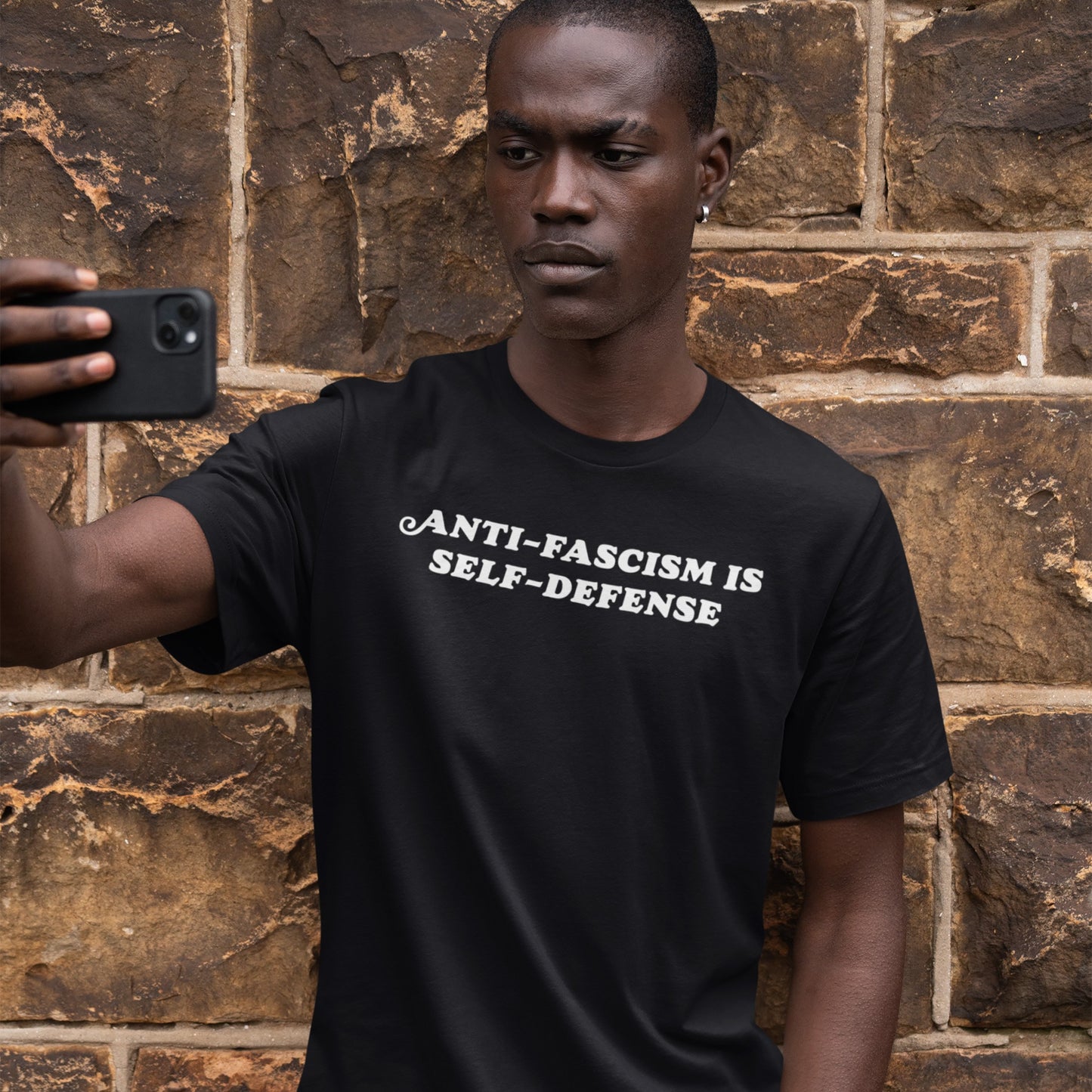 Young man holds a smartphone, wearing the Anti-Fascism Is Self-Defense T-shirt. The shirt features bold white lettering on black, embodying a clear, historical message of resistance and empowerment.