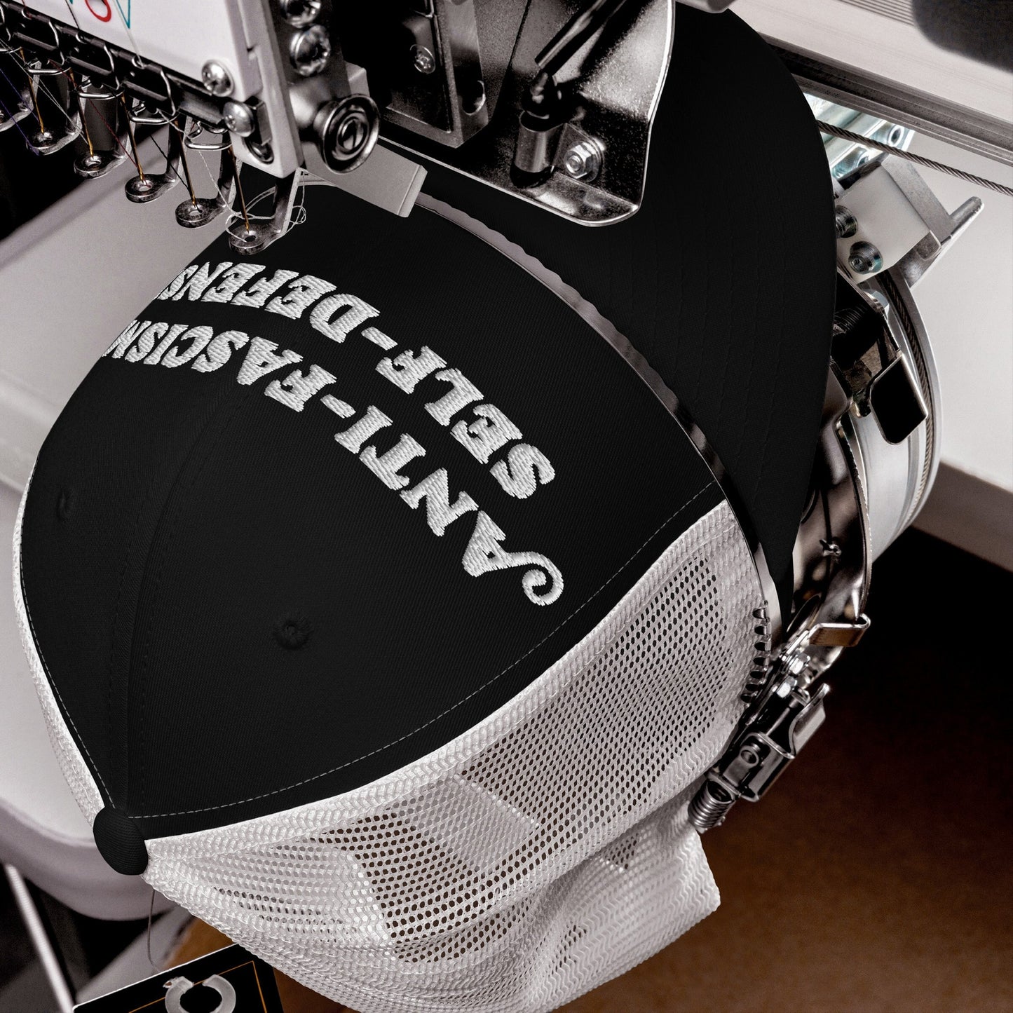 Black mesh-back trucker cap clamped in an embroidery machine, stitching Anti-Fascism Is Self-Defense in bold white script, embodying resistance.