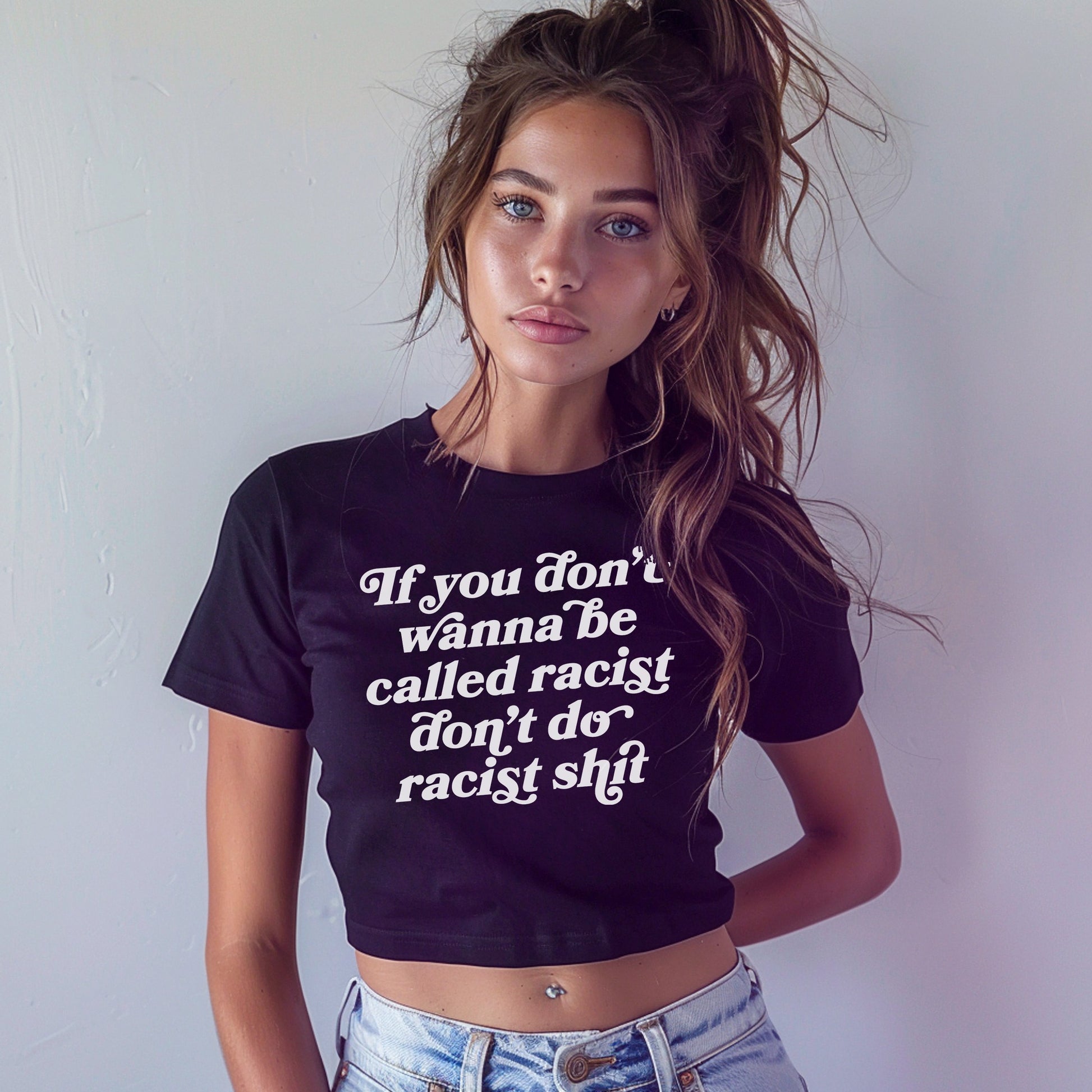 Young woman models the Anti-Racist Women's Baby Tee EU, featuring bold text on a black cropped T-shirt, paired with light-wash jeans. Her casual stance and piercing embody the tee's unapologetic message.