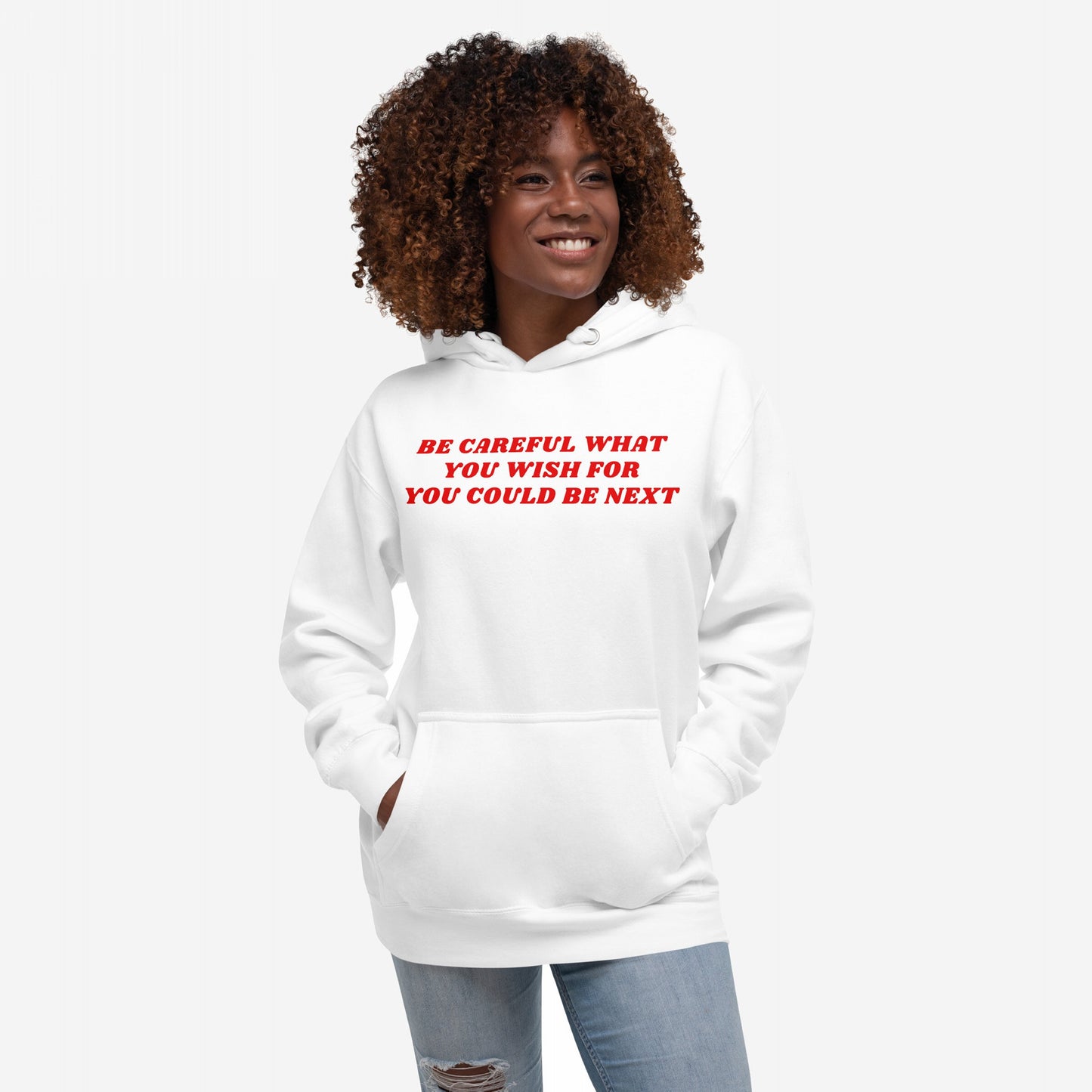 Young woman in a white Be Careful Hoodie & Sweatshirt with red slogan, hands in pockets, smiling. The hoodie challenges authoritarianism and supports equality, reflecting the store's rebellious ethos.
