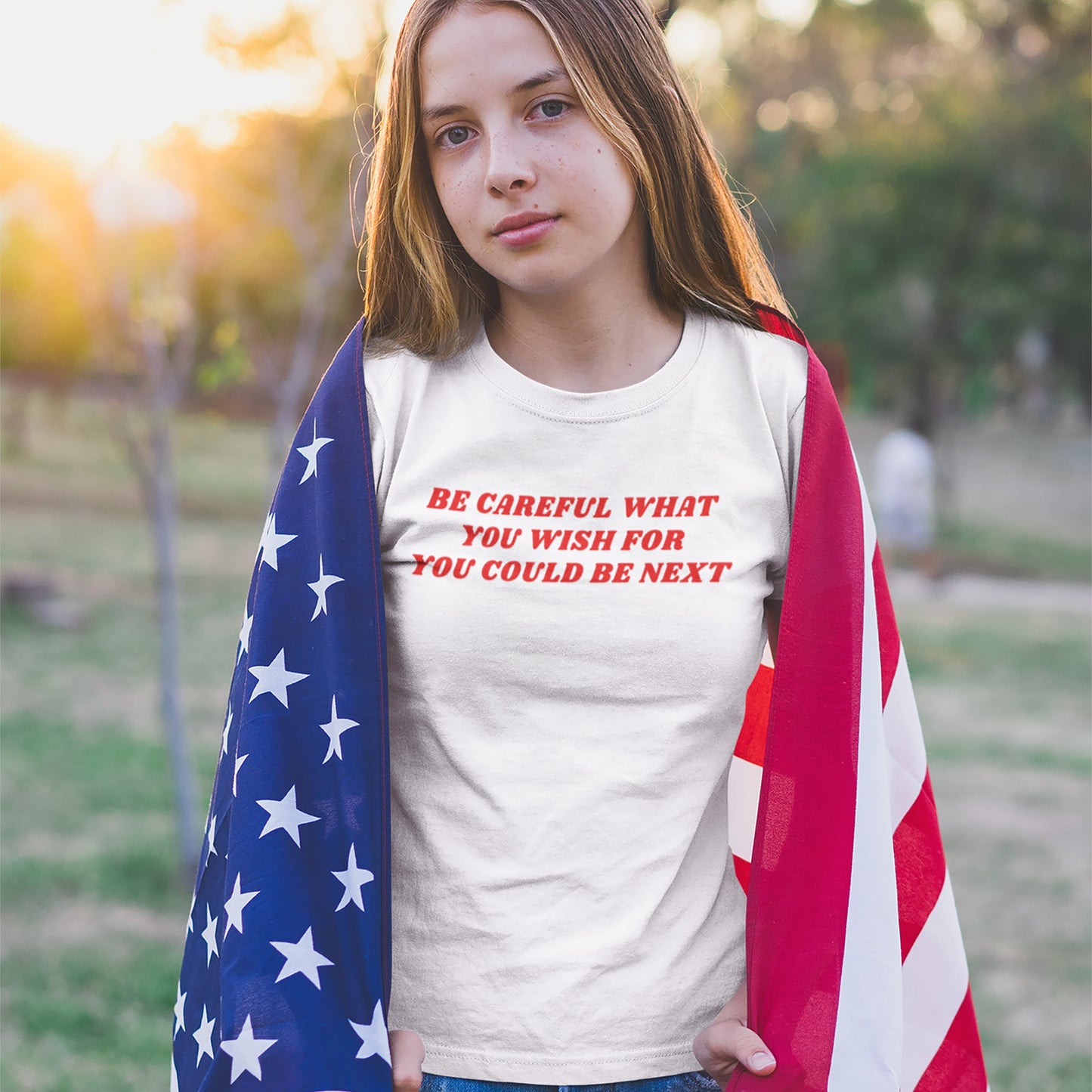 Young girl stands in a park at sunset wearing the Be Careful T-shirt, featuring bold red block letters challenging authoritarianism, draped in an American flag.
