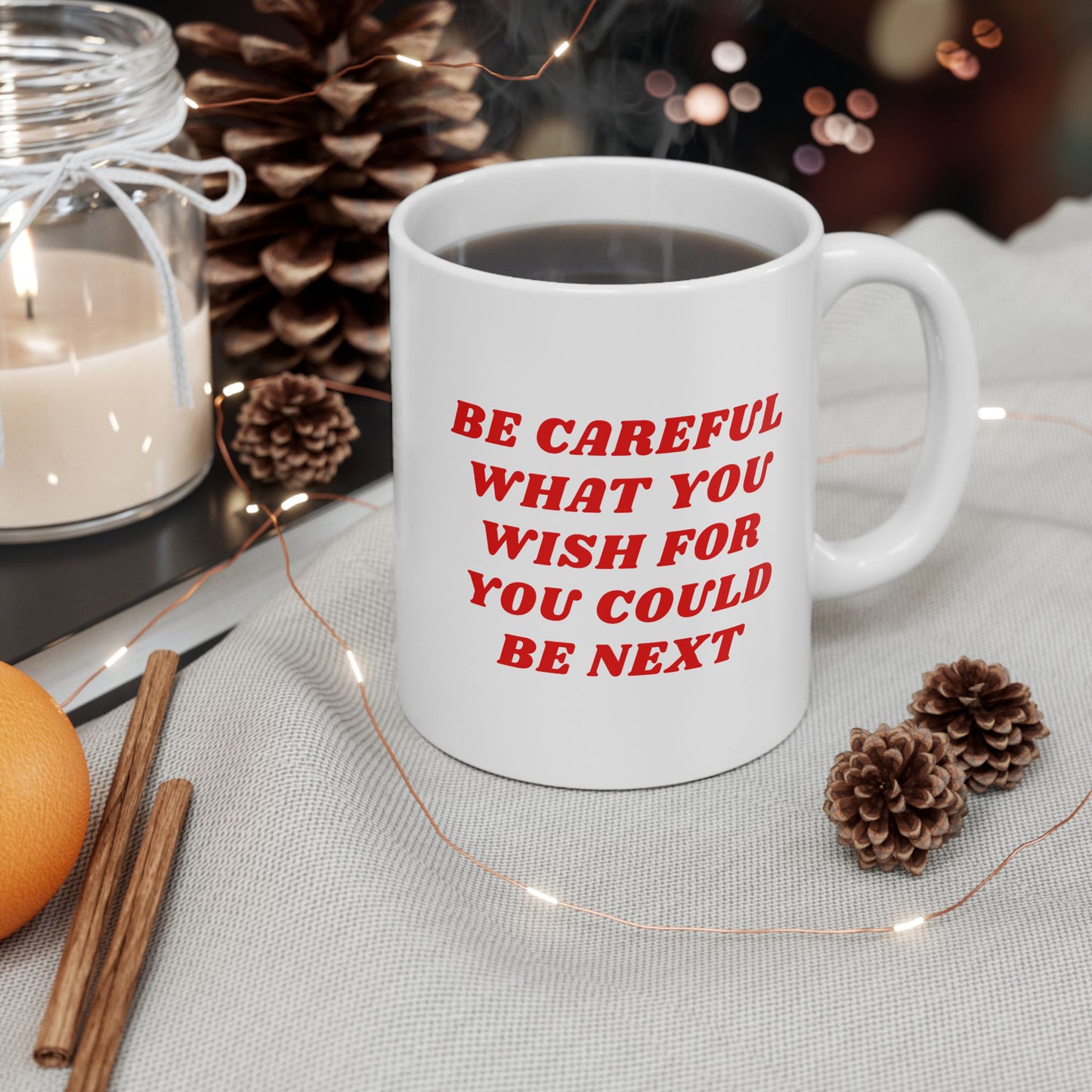 Cozy holiday scene with the Be Careful Mug featuring bold red text. Surrounded by a candle, pine cones, fairy lights, cinnamon sticks, and an orange on a soft fabric.