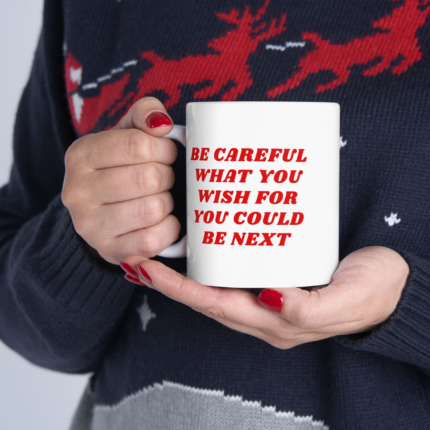 A person holds a Be Careful Mug, featuring bold red text warning, “BE CAREFUL WHAT YOU WISH FOR YOU COULD BE NEXT,” against a backdrop of a dark, festive-patterned sweater.
