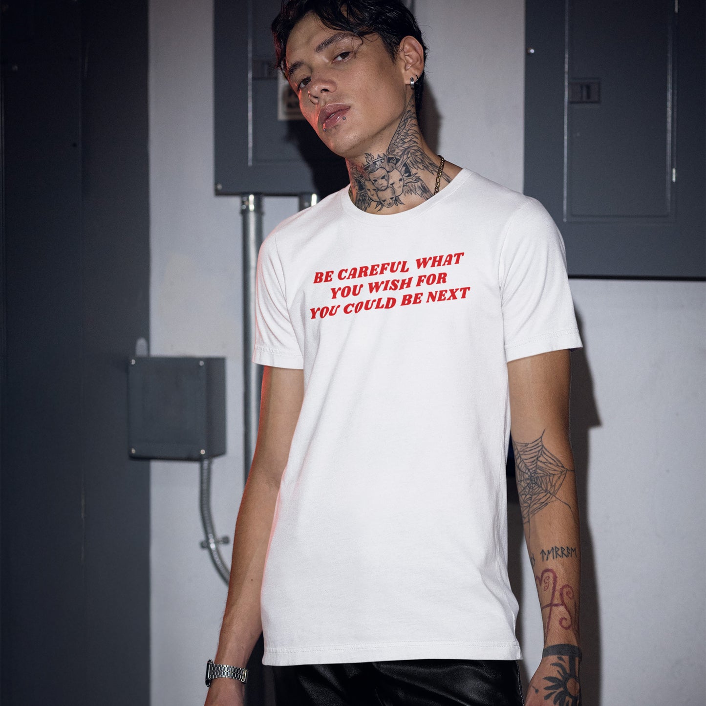 Young man wearing the Be Careful Shirt with bold red text, standing indoors with tattoos visible; the shirt delivers an anti-authoritarian message. Background features electrical panels; mood lighting emphasizes the design.