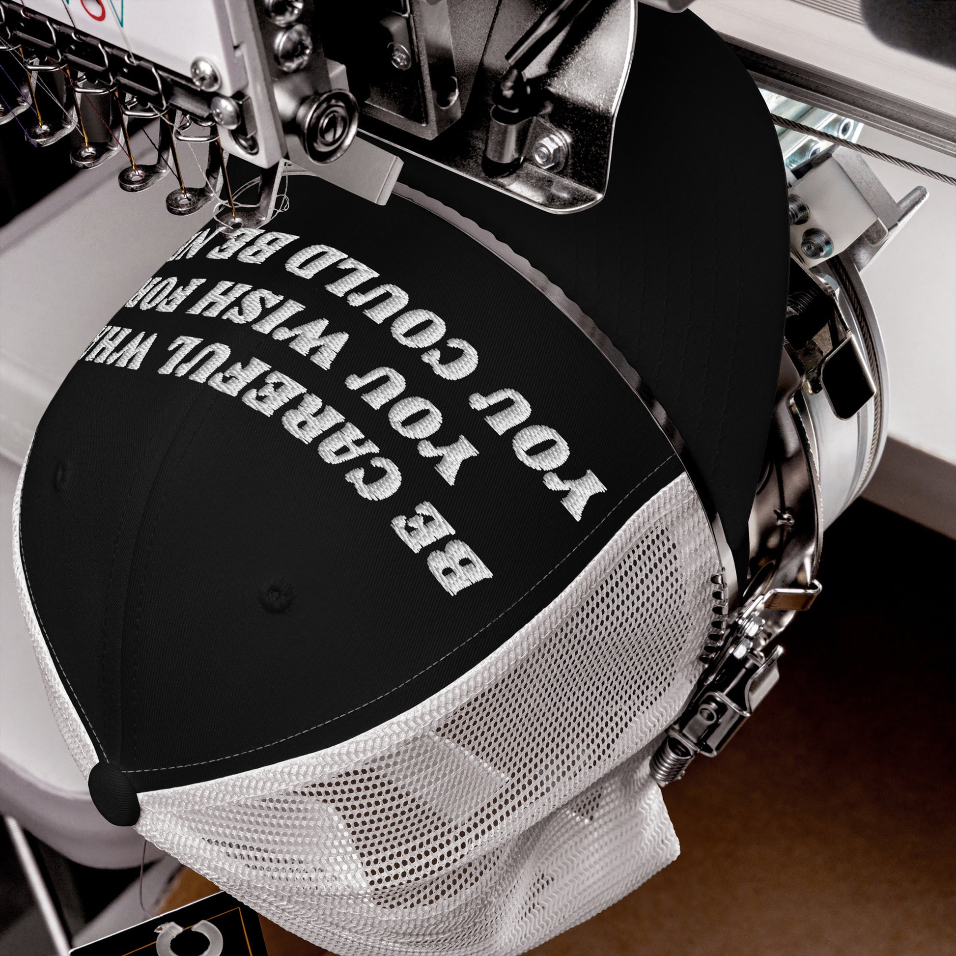 Black trucker-style cap with white mesh back on an embroidery machine, featuring the bold message Be Careful What You Wish For on the front, reflecting a strong progressive stance.