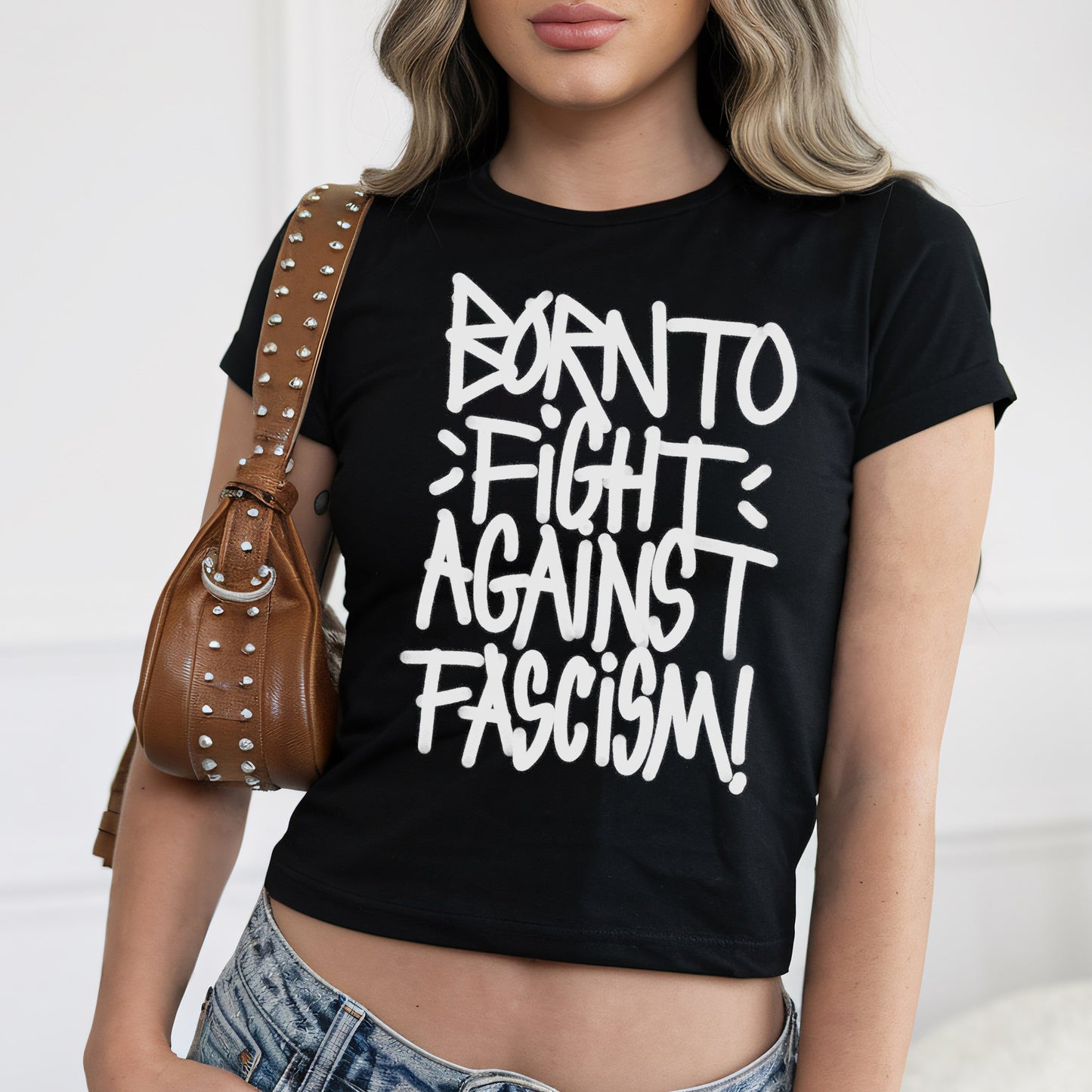 Born To Fight Against Fascism Graffiti Women's Baby Tee, featuring bold white lettering on black fabric, worn with light-wash jeans. The cropped design radiates defiance, epitomizing anti-fascist sentiment and empowerment.