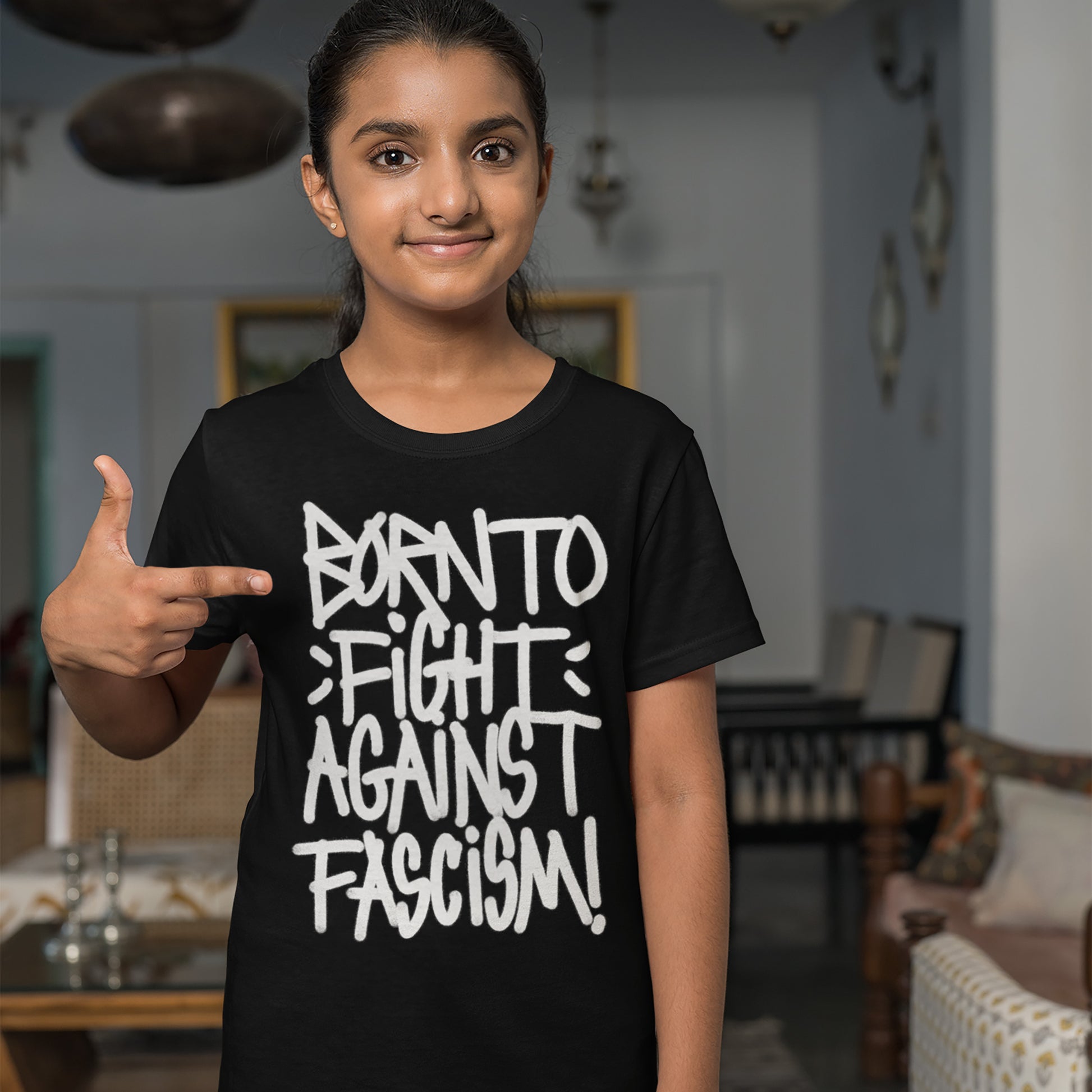 Young girl in a living room wearing the Born To Fight Against Fascism Graffiti Shirt, pointing at the bold, graffiti-style slogan on her black t-shirt, embodying resistance and defiance.