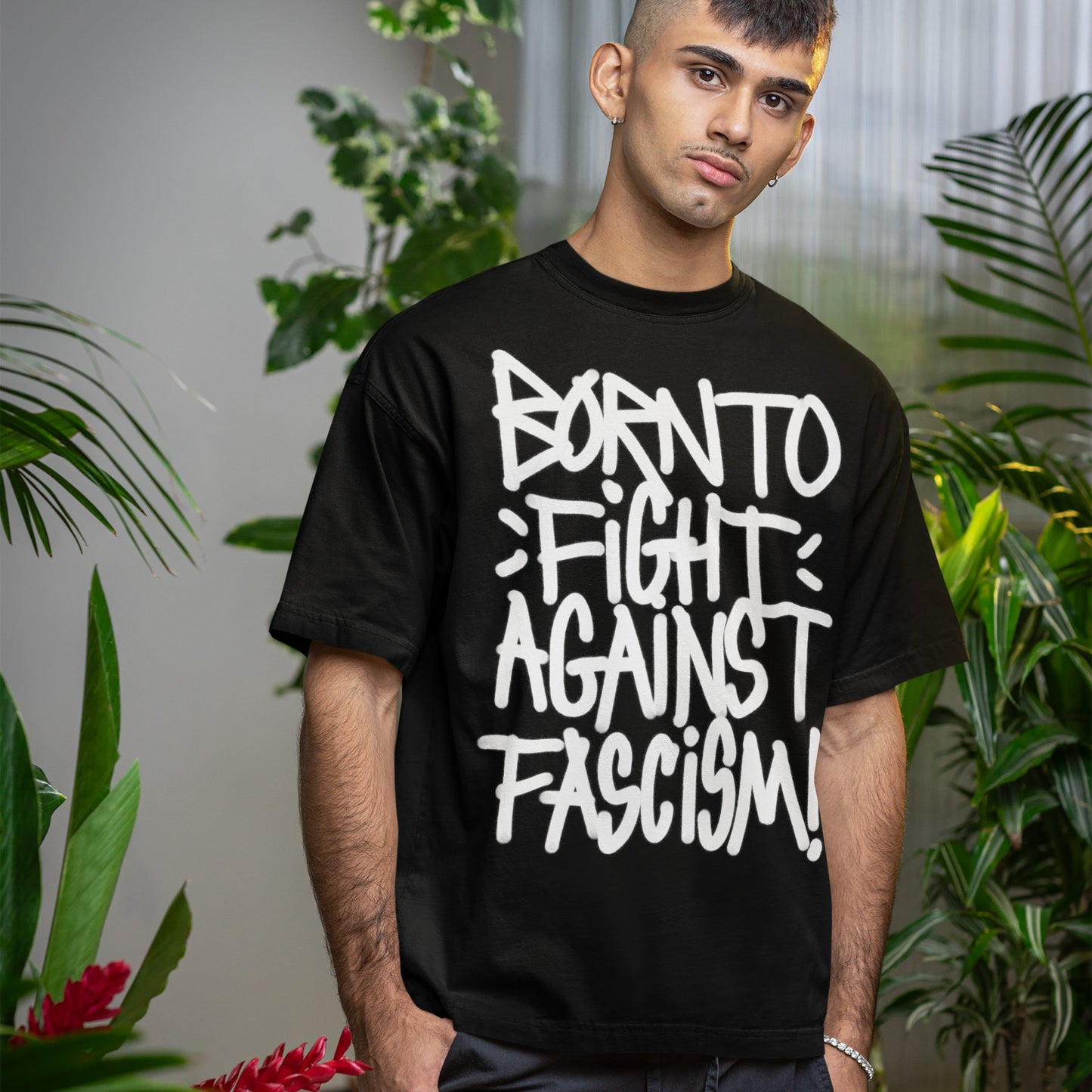 Young man wearing the Born To Fight Against Fascism Graffiti T-shirt, featuring bold white graffiti-style text. He stands amidst leafy plants, embodying the shirt's activist spirit with a neutral expression.