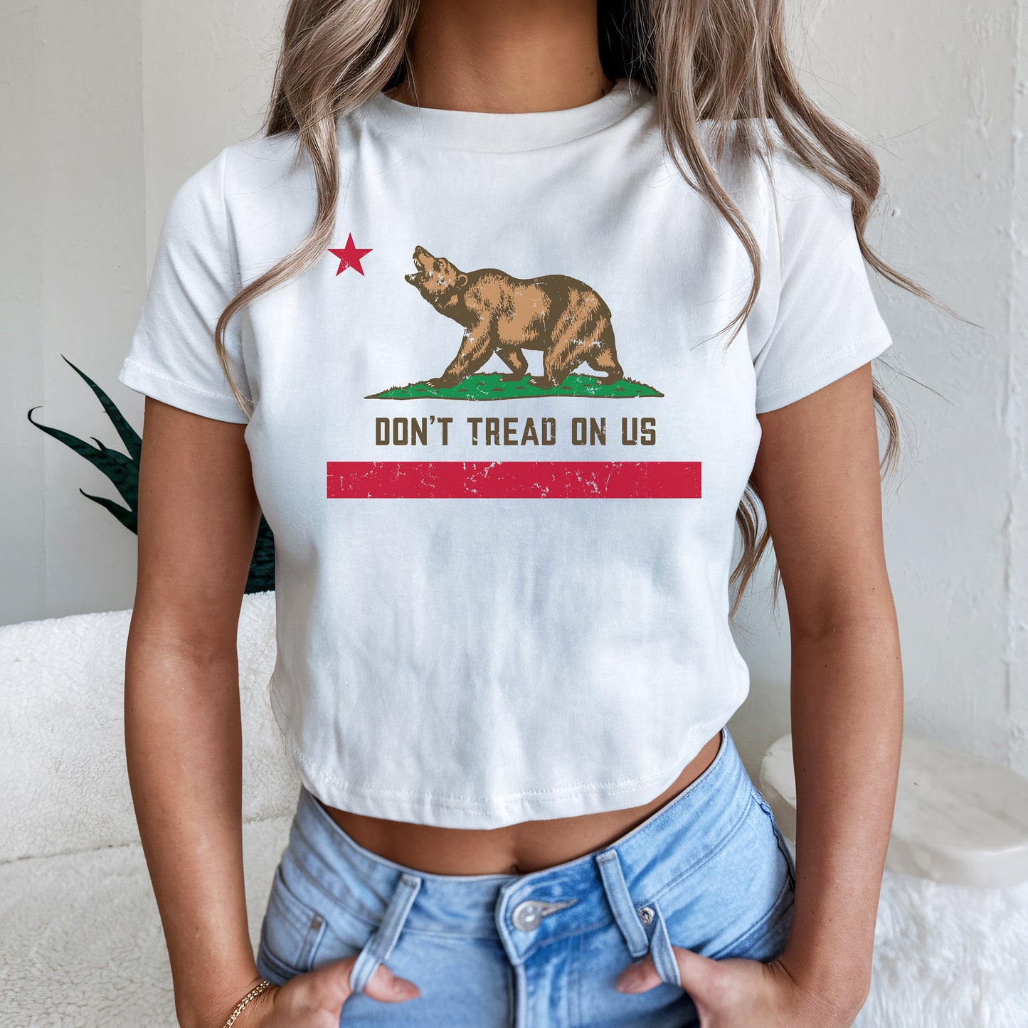 California Gadsden Flag Women's Baby Tee features a stylized bear and star with DON’T TREAD ON US slogan, worn with thumbs in jeans pockets, expressing bold resistance and state pride.