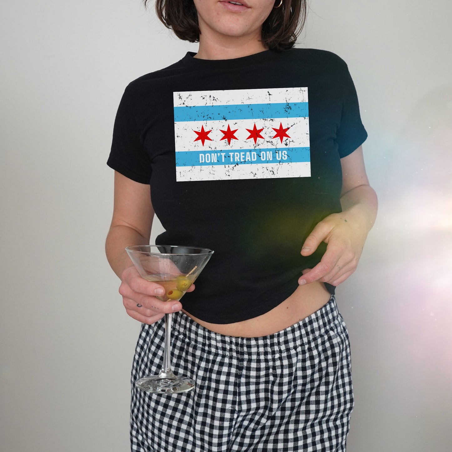 Chicago Gadsden Flag Women's Baby Tee featuring a cropped design with Chicago flag-inspired protest print, held by a person with gingham bottoms and a martini glass, embodying bold, stylish activism.