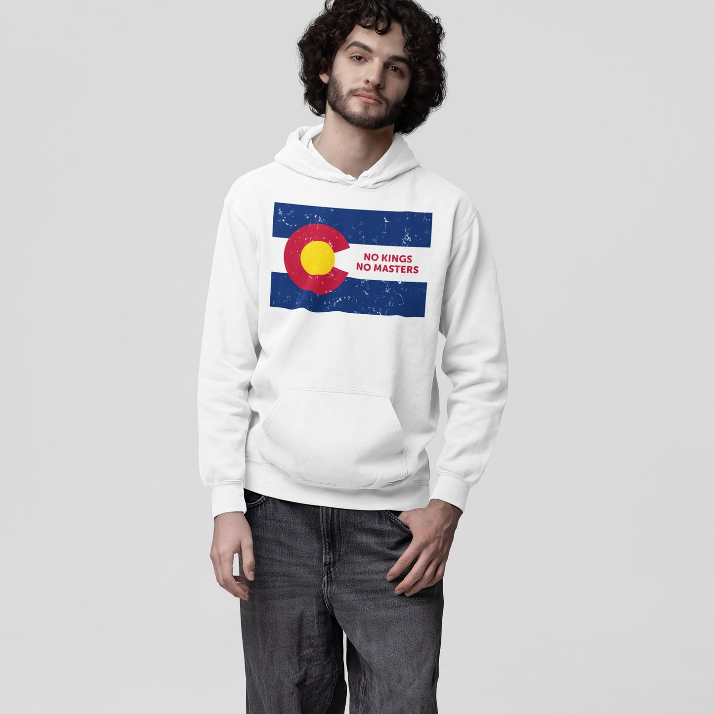 Young man wearing a white hoodie with a distressed Colorado flag and NO KINGS NO MASTERS slogan, promoting autonomy and rebellion. The Colorado No Kings Flag Shirt supports the Colorado Immigrant Rights Coalition.