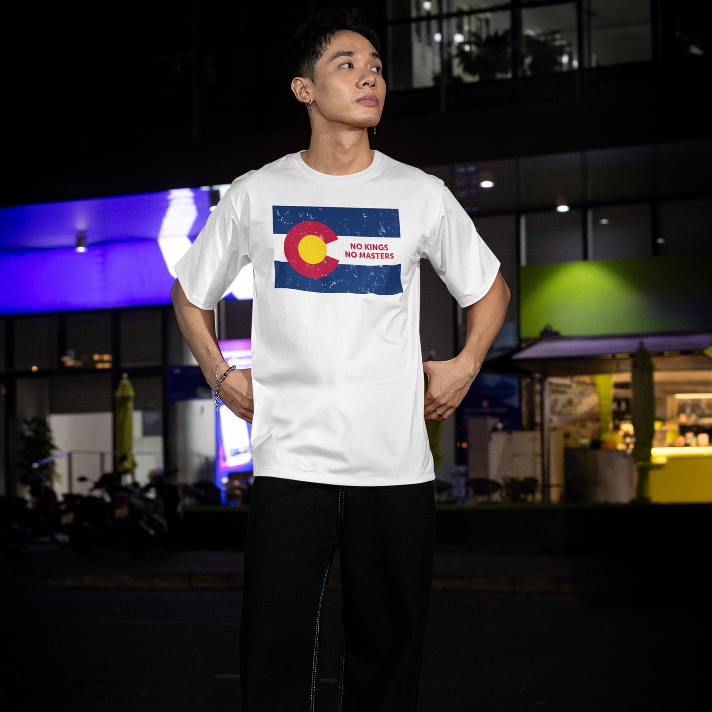 Young person wearing the Colorado No Kings Flag Shirt featuring a distressed Colorado-flag motif with “NO KINGS NO MASTERS” slogan, standing in front of a lit-up urban storefront at night.