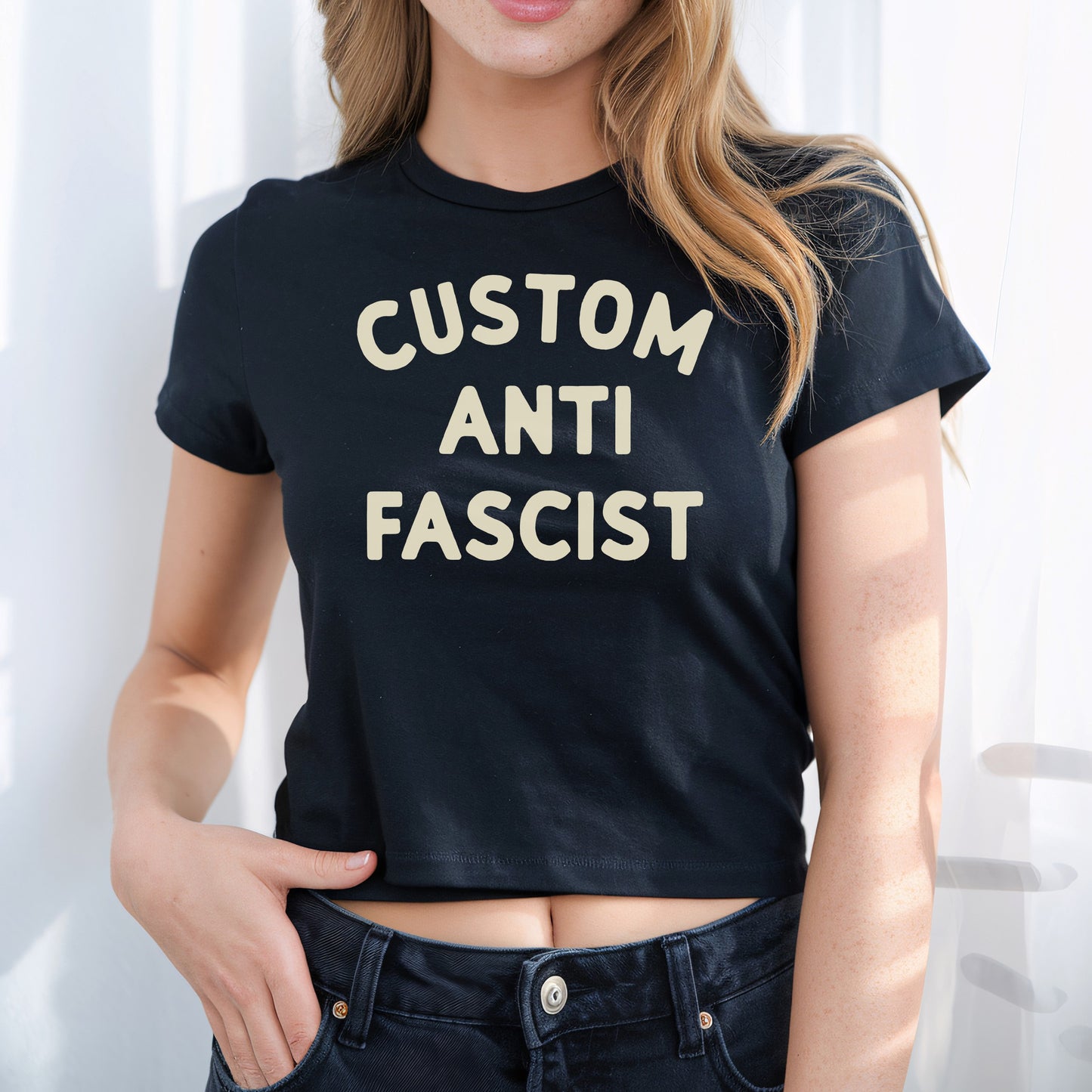 Young person wearing the Custom Anti-Fascist Women's Baby Tee, featuring bold cream text. This cropped black T-shirt reflects a retro Y2K style, paired with dark denim jeans, embodying activism and individuality.