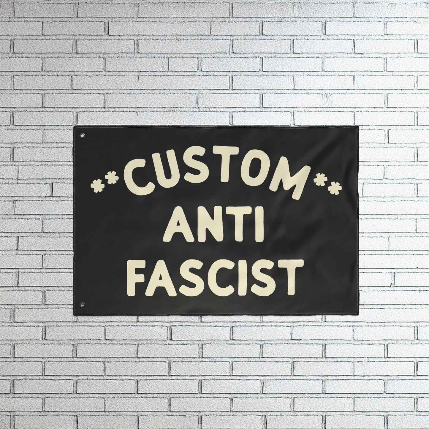 Custom Anti-Fascist Flag with bold off-white lettering on a black fabric, mounted on a white brick wall. This customizable flag powerfully communicates anti-fascist sentiment, ideal for rallies or home display.
