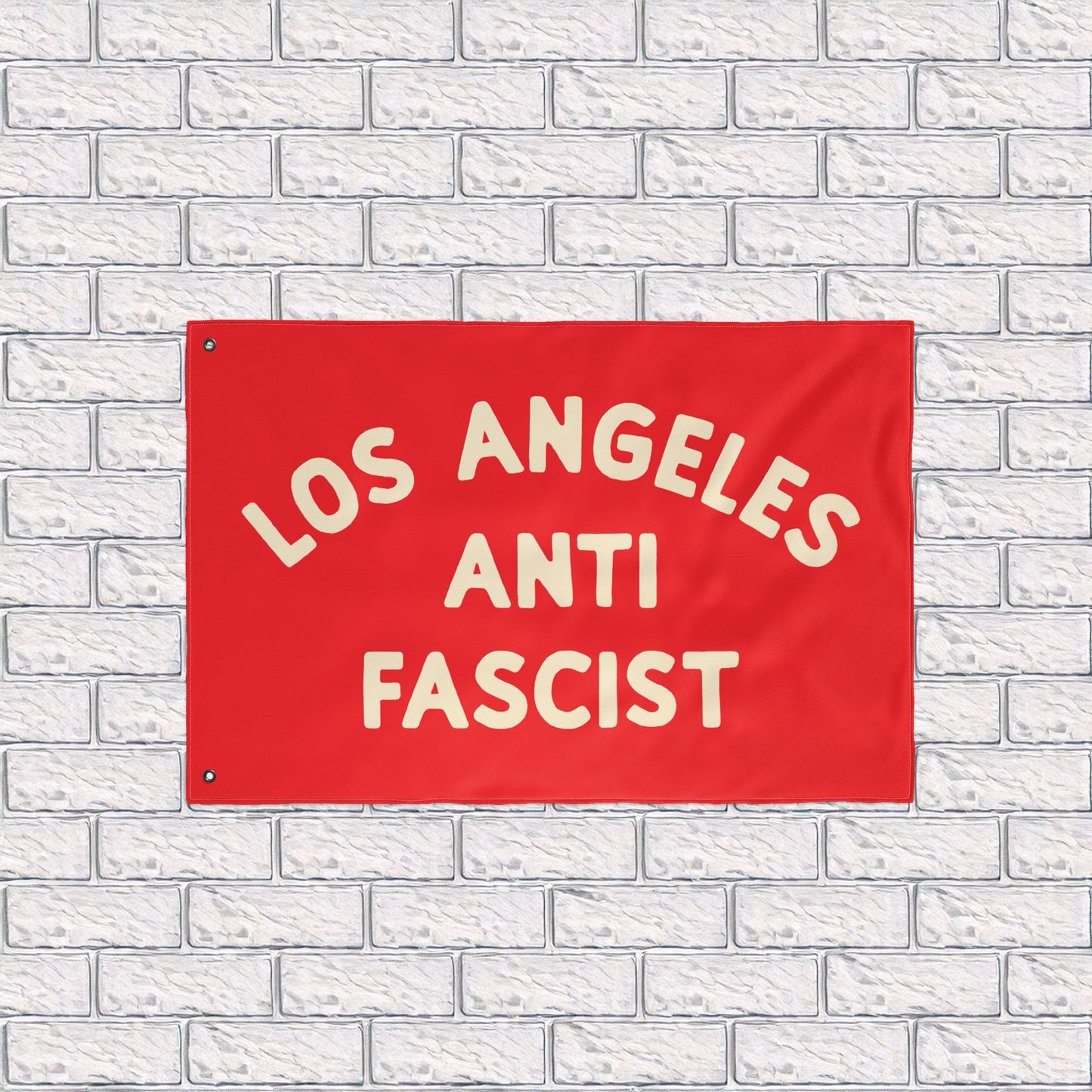 Custom Anti-Fascist Flag featuring LOS ANGELES ANTI FASCIST in bold cream text on a red background, mounted against a white brick wall. Durable polyester with metal grommets, ideal for rallies or home display.