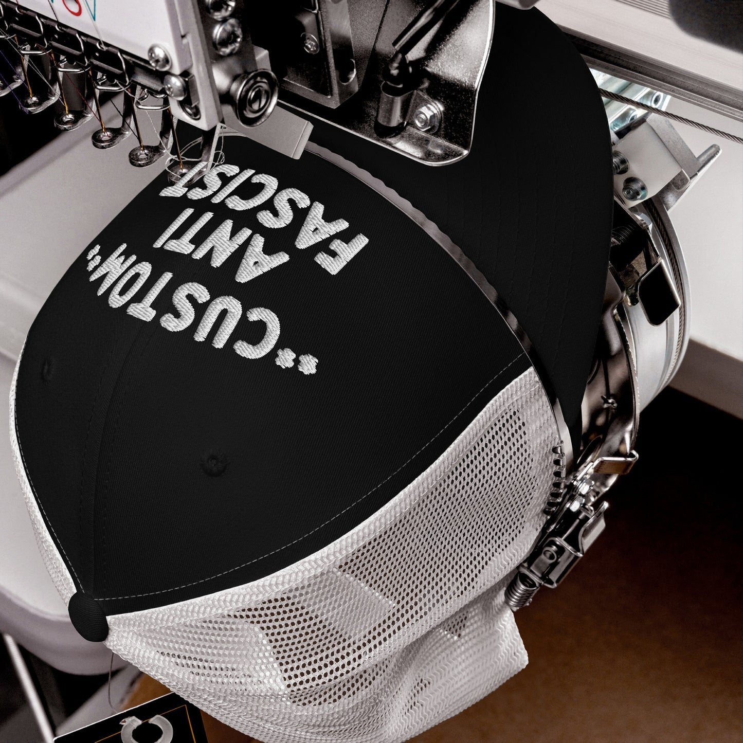 Custom Anti-Fascist Trucker Hat in embroidery machine, stitching white thread on black front panel with white mesh back. Personalize your message, showcasing bold anti-fascist stance with breathable design.