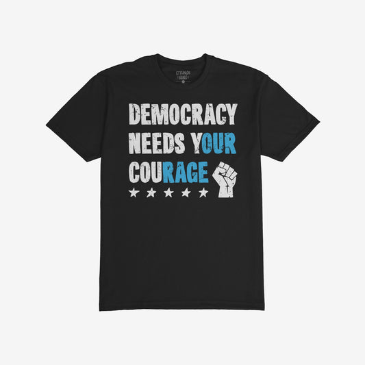 Democracy T-shirt features DEMOCRACY NEEDS YOUR COURAGE in distressed lettering with stars and a raised fist, embodying a bold statement of resistance and solidarity across borders.