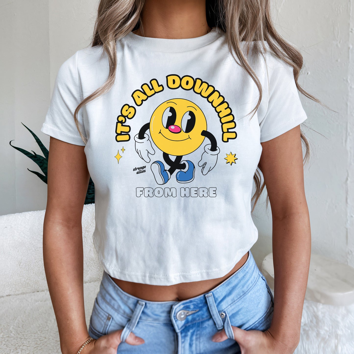 Woman models the Downhill Women's Baby Tee, featuring a cartoonish yellow smiley with limbs, exuding playful existentialism with the slogan “IT’S ALL DOWNHILL FROM HERE,” paired with light-wash jeans.