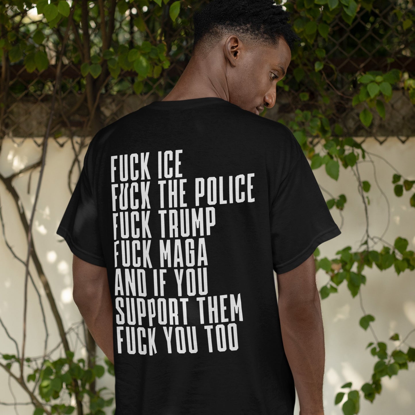 Person wearing the Fuck 'Em All Back Print T-shirt, featuring bold text denouncing ICE, police, Trump, and their supporters, standing by a fence with foliage.